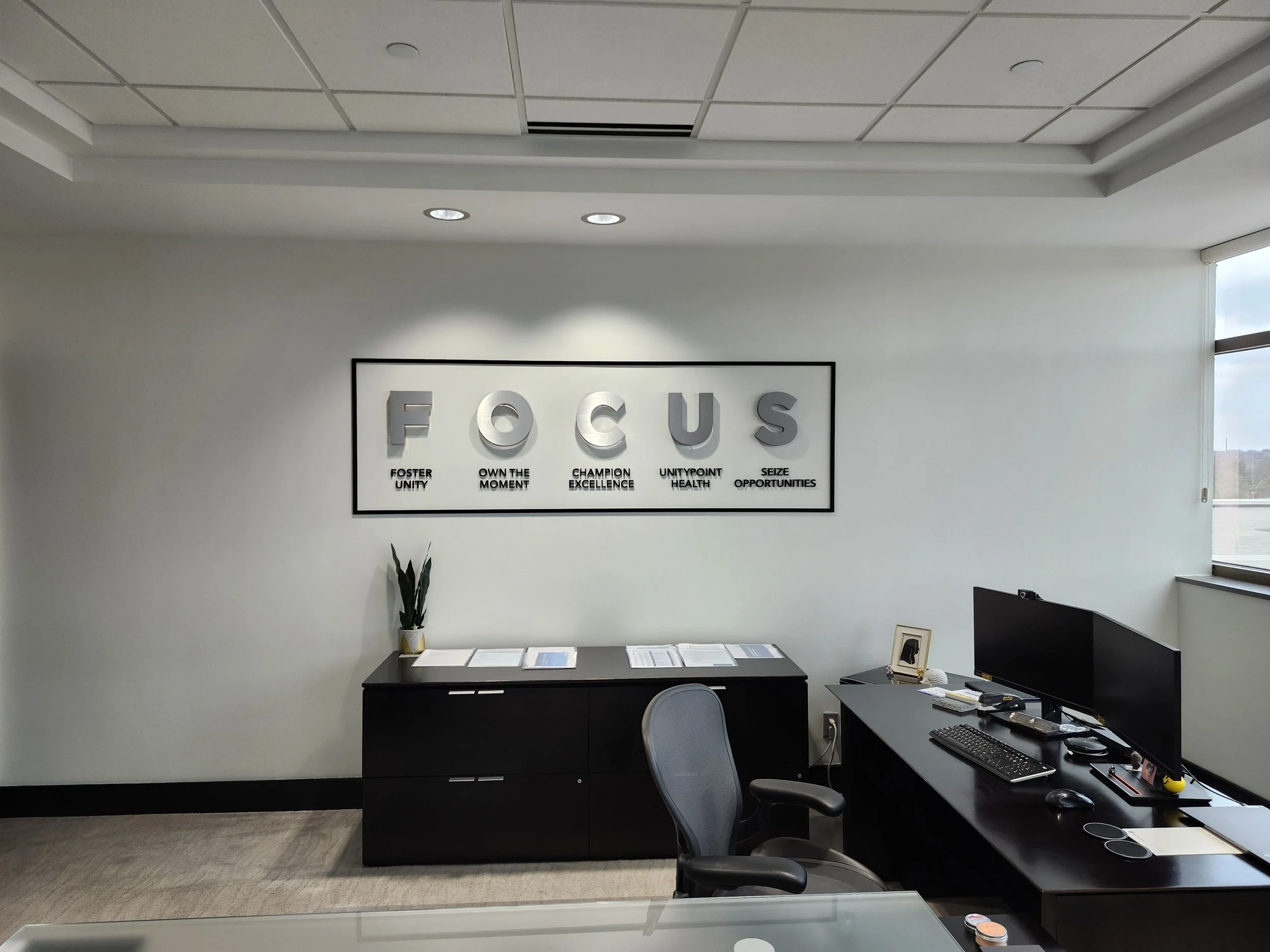 An office with a black desk and chair, a computer monitor, a keyboard, and office supplies. The wall features a poster with the word 'FOCUS' and motivational phrases underneath. There is a window on the right side letting in natural light.