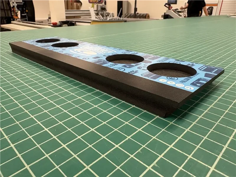 A black rectangular piece of foam with four circular cutouts on a green cutting mat, with a background of a workspace with tools and equipment.