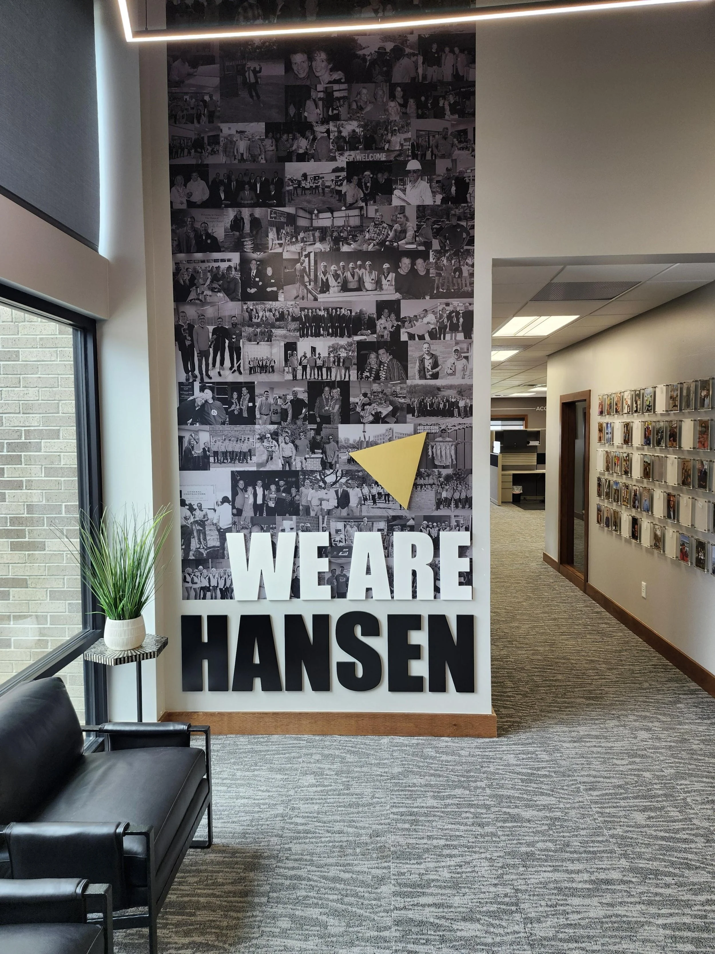 A photo of an office lobby area with a large black and white collage wall vinyl featuring pictures of people, a gold metal triangle, and the words 'WE ARE HANSEN' in large white and black acrylic dimensional letters in the foreground. There is a blac