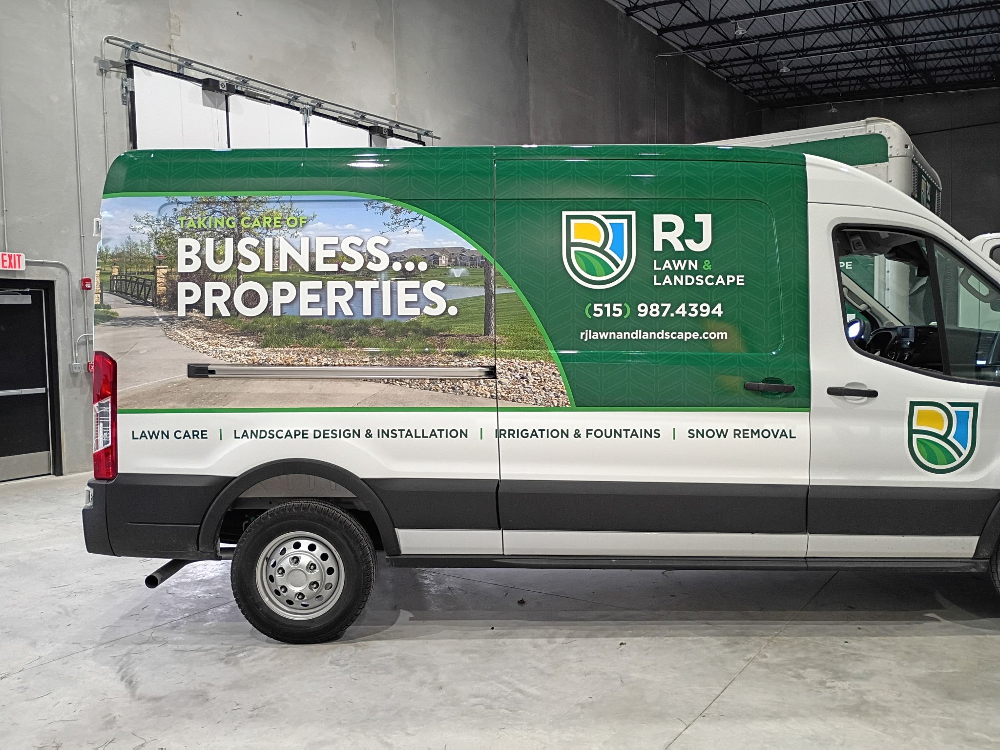 A white work van with green and blue advertising for RJ Lawn & Landscape, displaying services like lawn care, landscaping, irrigation, fountains, and snow removal, parked inside a warehouse or garage.