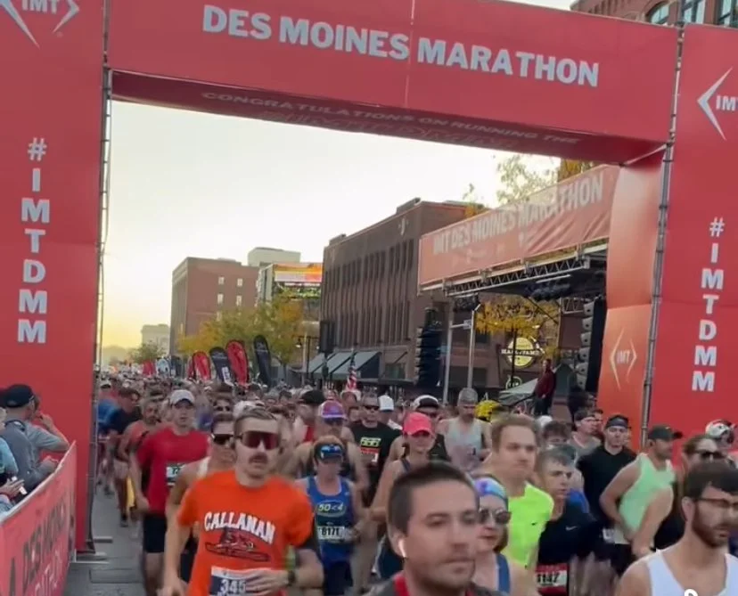 Starting line of the Des Moines Marathon with runners, a large red archway, and city buildings in the background.