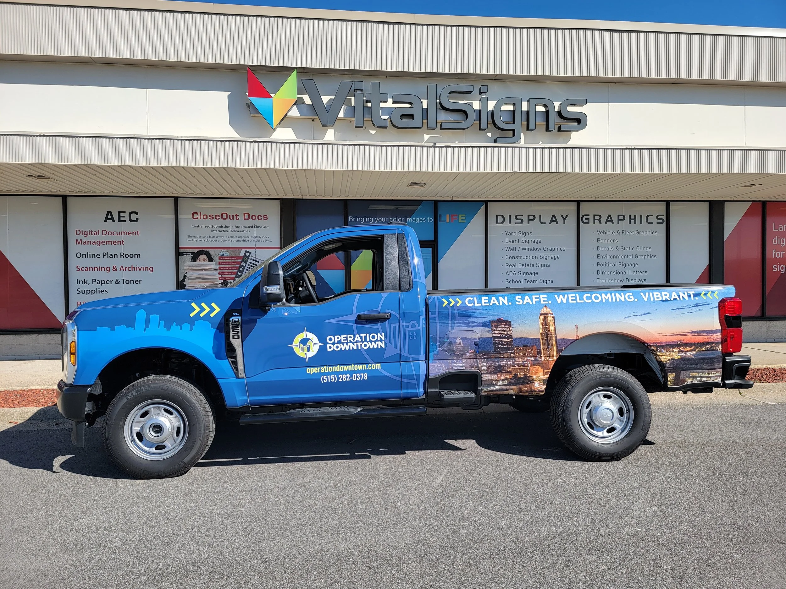 A blue truck parked in front of a building with signage for Vital Signs and Operation Downtown, with a city skyline graphic on the side of the truck and the slogan 'CLEAN. SAFE. WELCOMING. VIBRANT.'