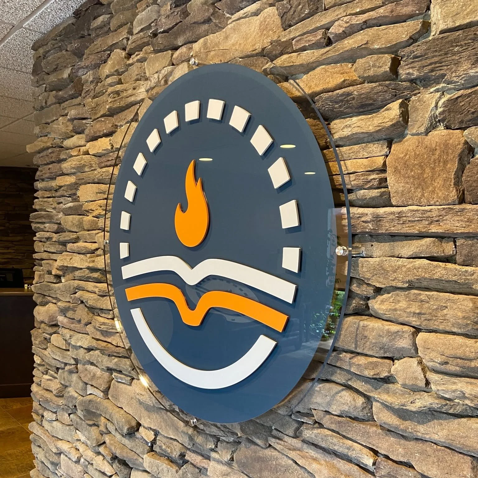 A wall-mounted acrylic sign with dimensional acrylic pieces in branded colors on front , and reverse printed background branded color. The large acrylic backer has polished edges and is mounted with standoffs against a stone wall.