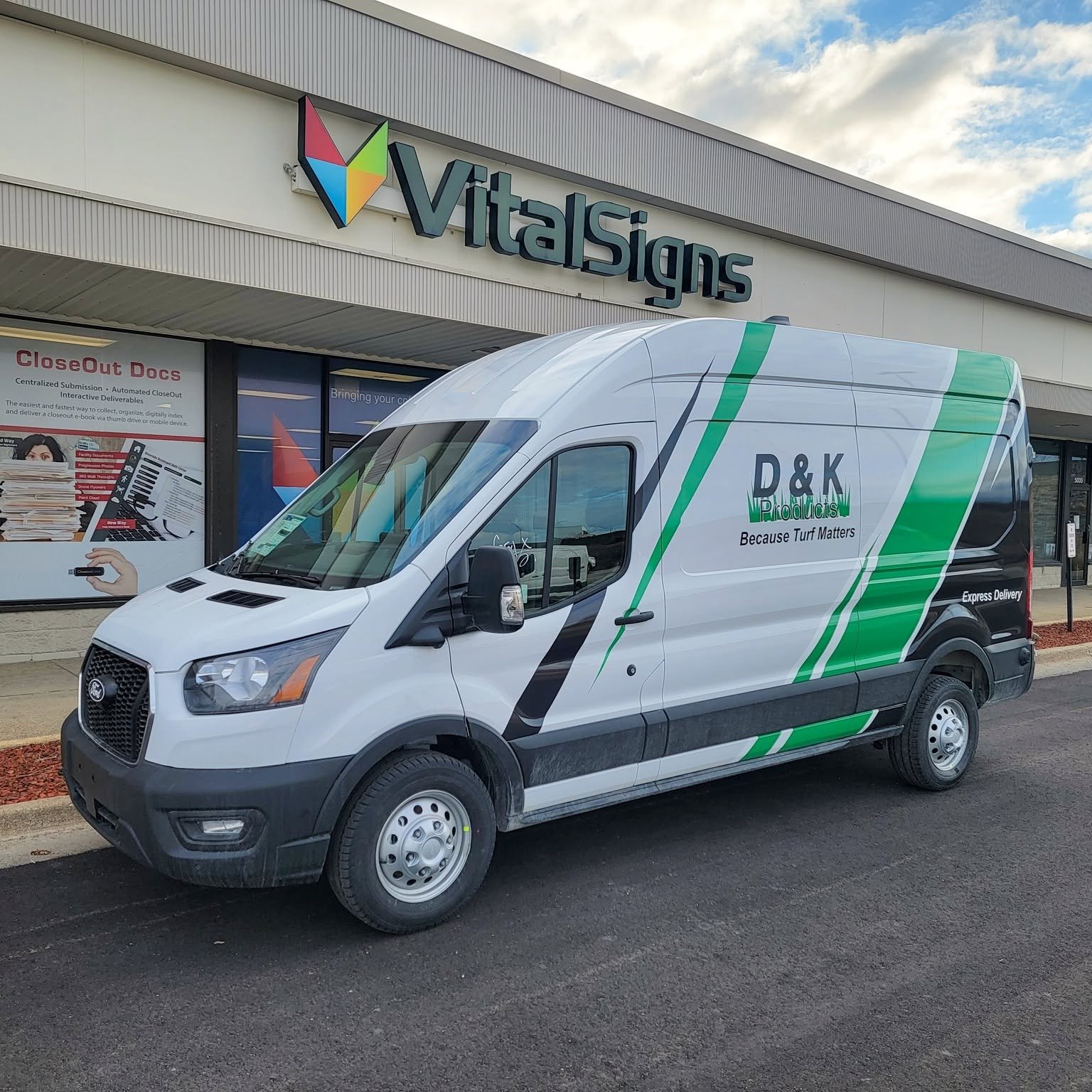 A white delivery van with green and black stripes parked in front of a Vital Signs store. The van has the logo 'D&K Products' and the slogan 'Because Turf Matters' on its side. The store's sign is visible on the building behind the van, along with a 