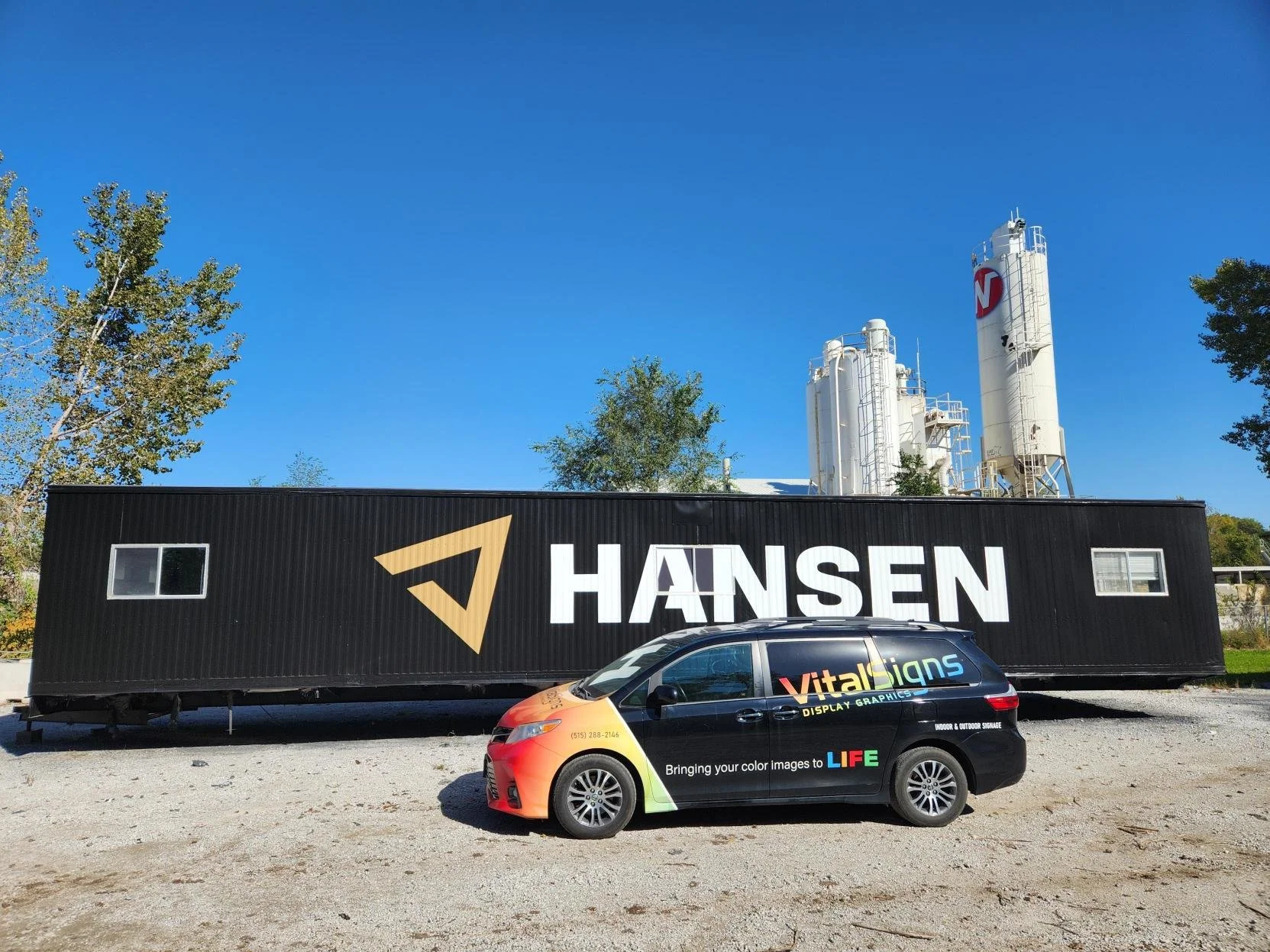 A black trailer with the logo and name of Hansen displayed, parked outside near industrial silos. In front of the trailer, a black van with colorful graphics and the phrase "Bringing your color images to LIFE" is parked.