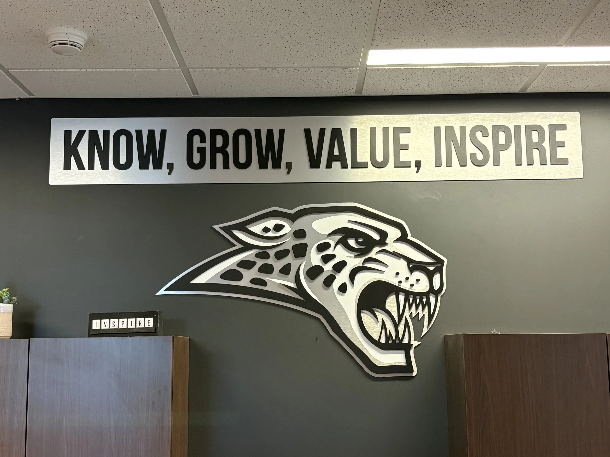 Interior of a building with a metallic sign that reads 'KNOW, GROW, VALUE, INSPIRE' on the wall. Below the sign is a portion of a stylized animal logo, possibly a bear. The ceiling has a fire sprinkler and fluorescent lights.