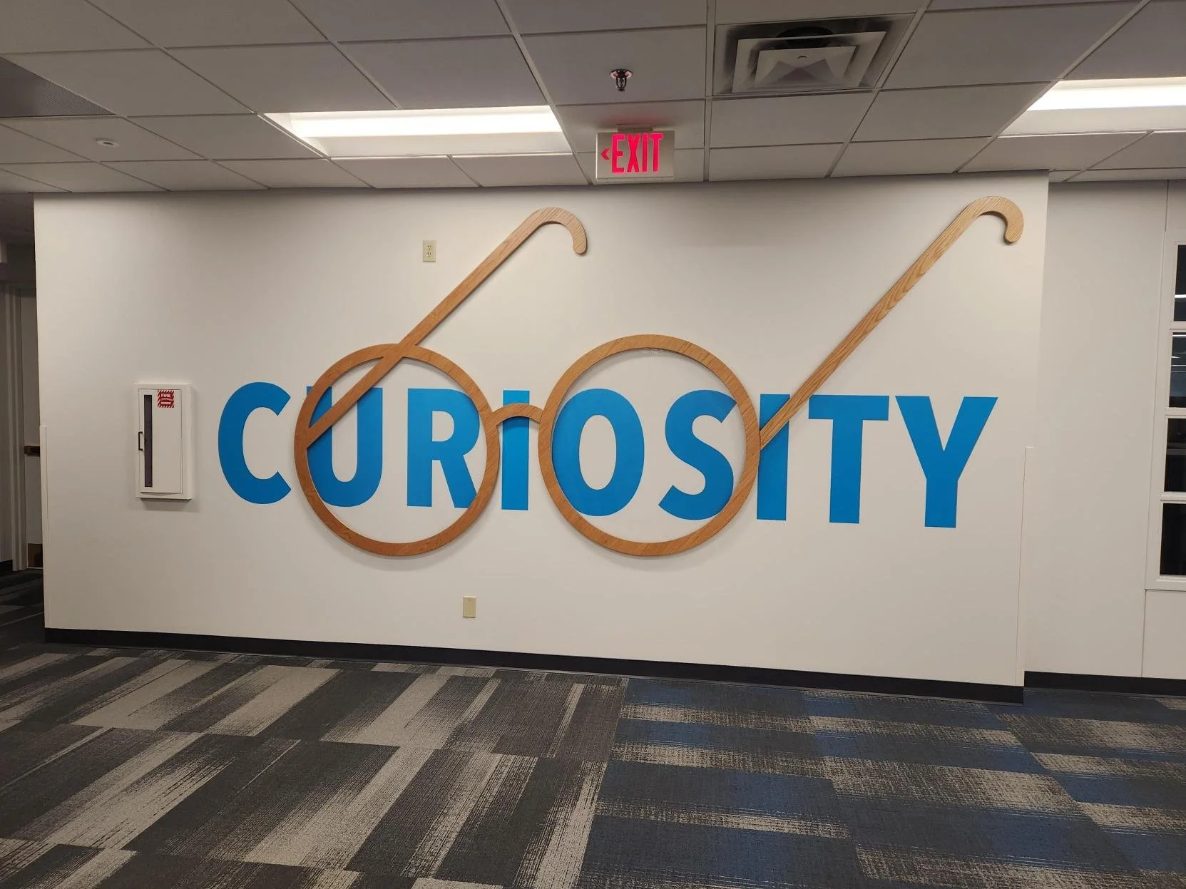 Wall decoration with the word 'CURIOSITY' in large blue letters, featuring a stylized magnifying glass with two wooden rims and handles, symbolizing curiosity and exploration, inside a room with a ceiling air vent, a red exit sign, and a fire alarm o