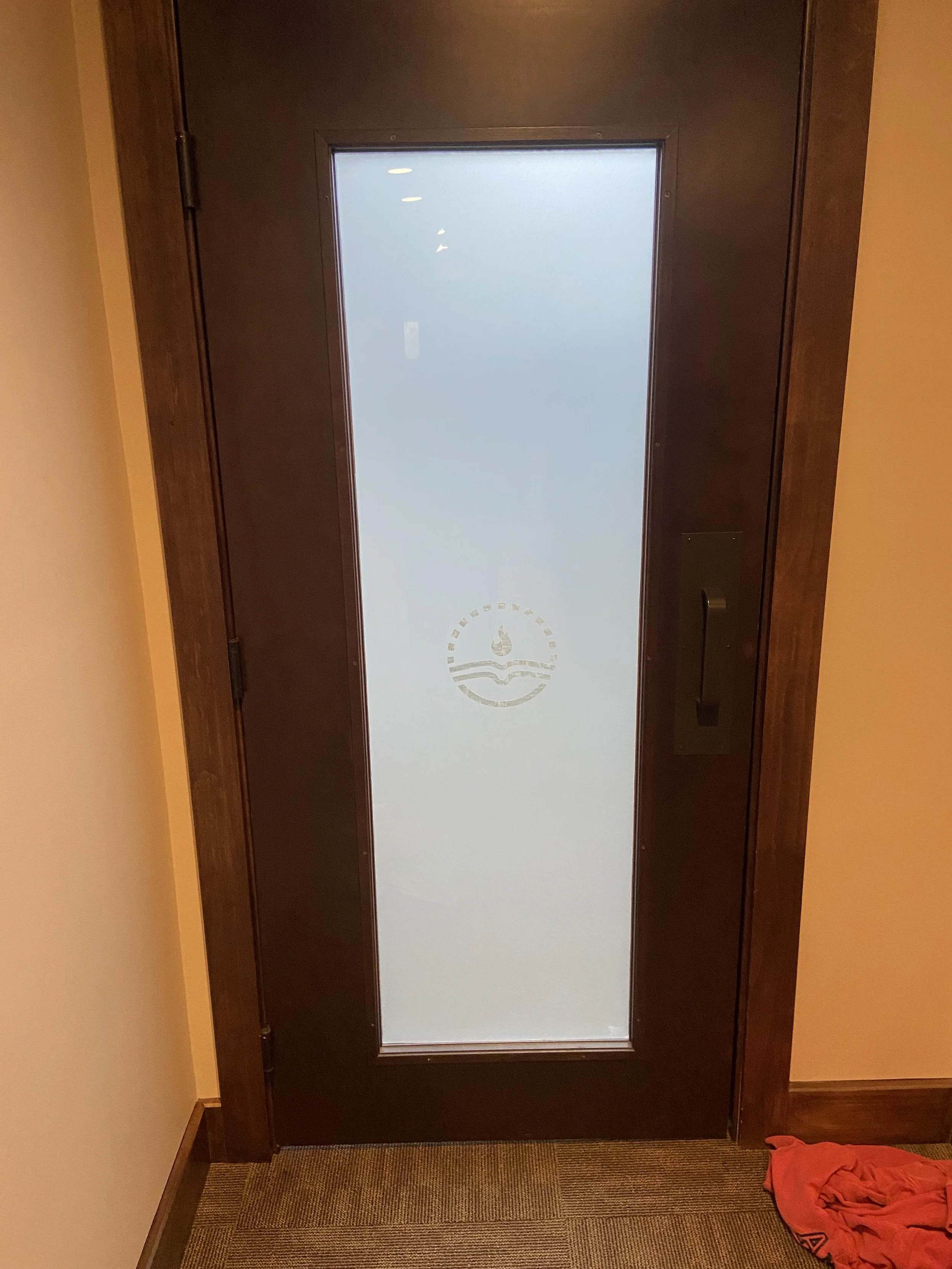 A door frosted with alogo at the bottom center, and the frame is made of dark wood. The elevator is located in a hallway with beige walls and a carpeted floor. There is an orange cloth or towel on the floor to the bottom right.