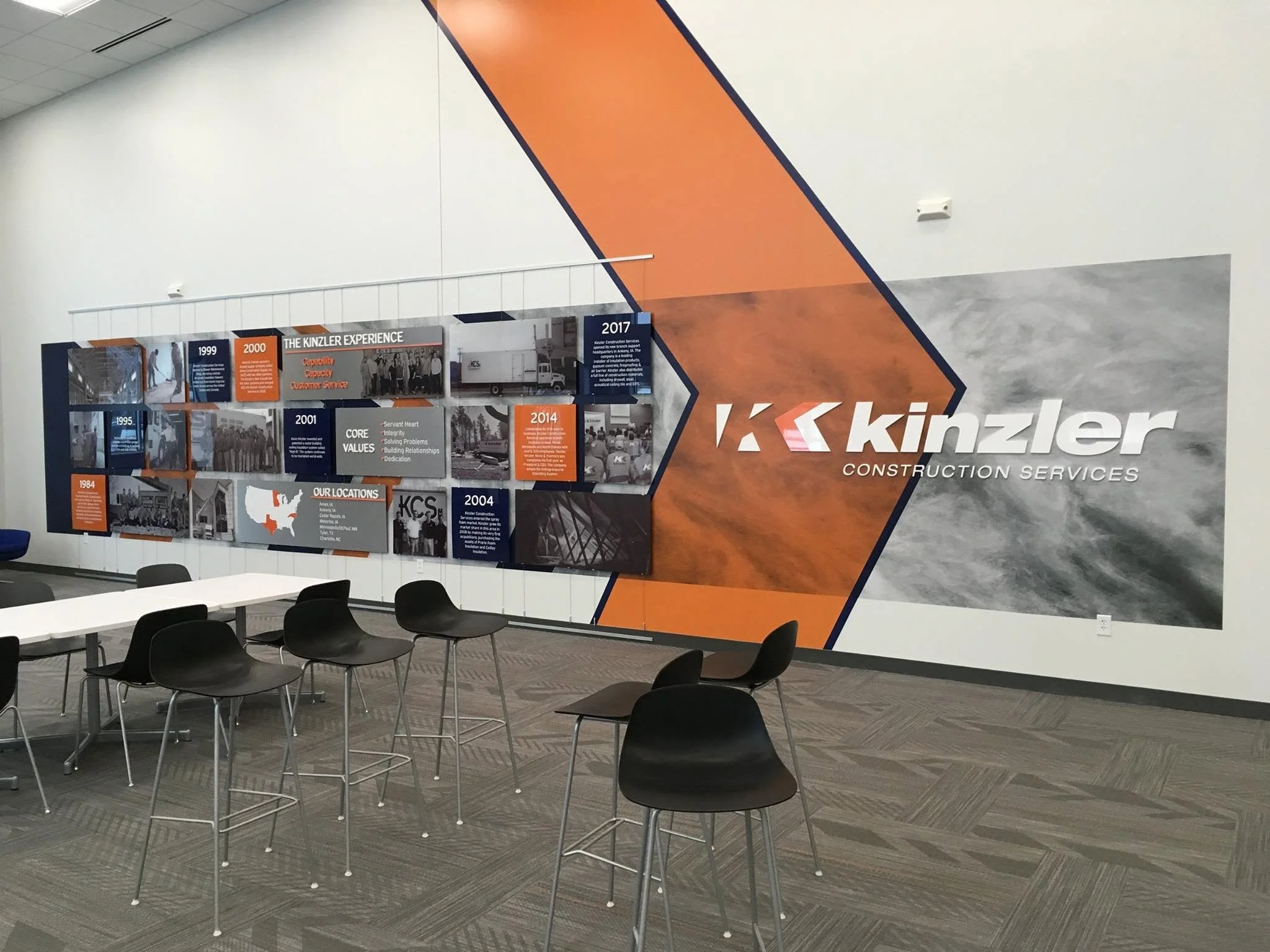 A conference room with a large wall display showing a timeline of Kinzler Construction Services' history from 1984 to 2017. The display features images, text, and branding, with modern furniture including chairs and tables.