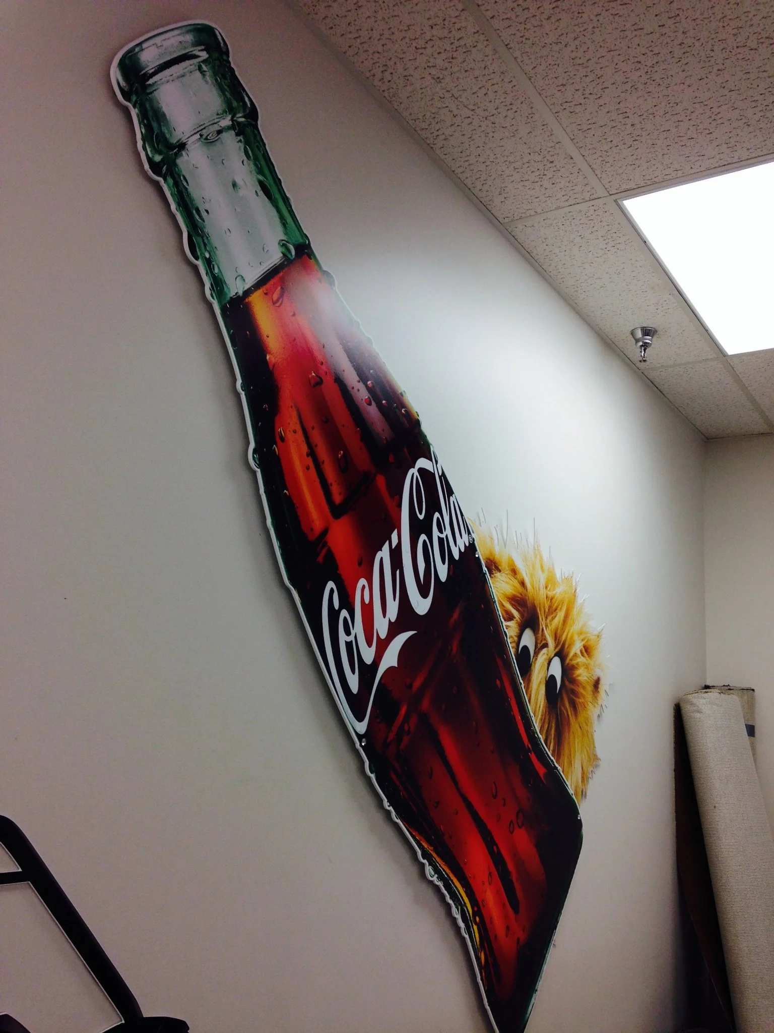 A large Coca-Cola sign featuring a dog with a lion costume on the right side of a plain light-colored wall, photographed at an angle.