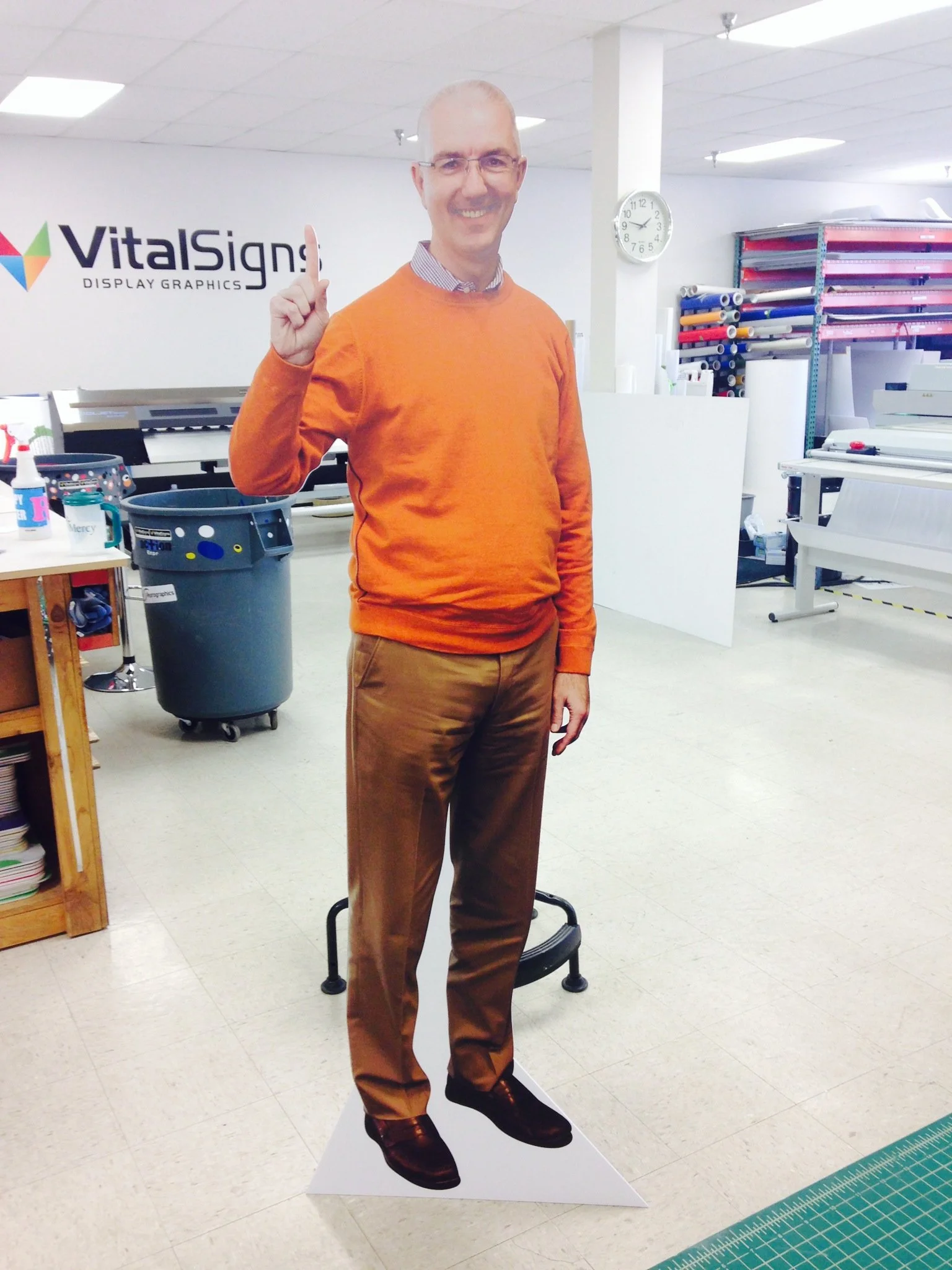 Full-length display cutout of a smiling man wearing glasses, an orange sweater, and brown pants, standing on a white triangular base in an office or workspace with shelves of rolls and a wall clock in the background.