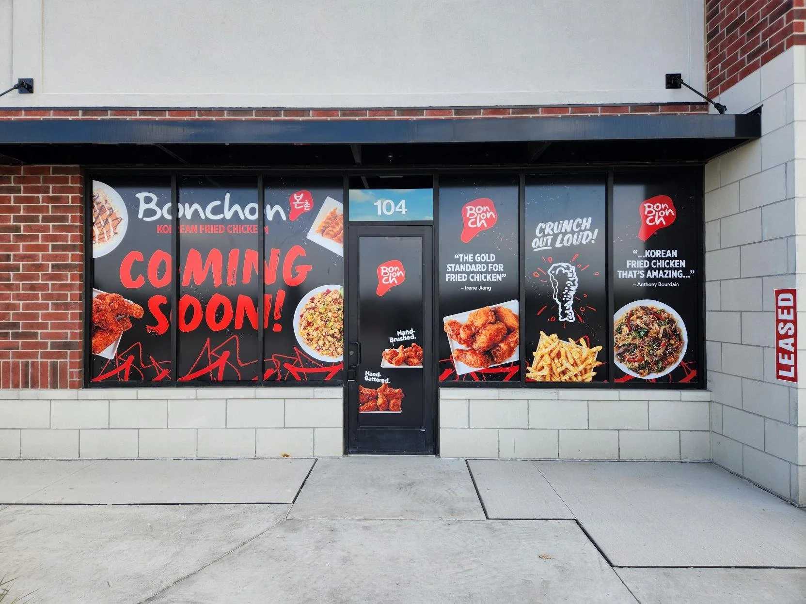 Storefront window advertising Korean fried chicken and meals, with text announcing upcoming opening and quotes about fried chicken, featuring images of fried chicken, noodles, and other dishes.