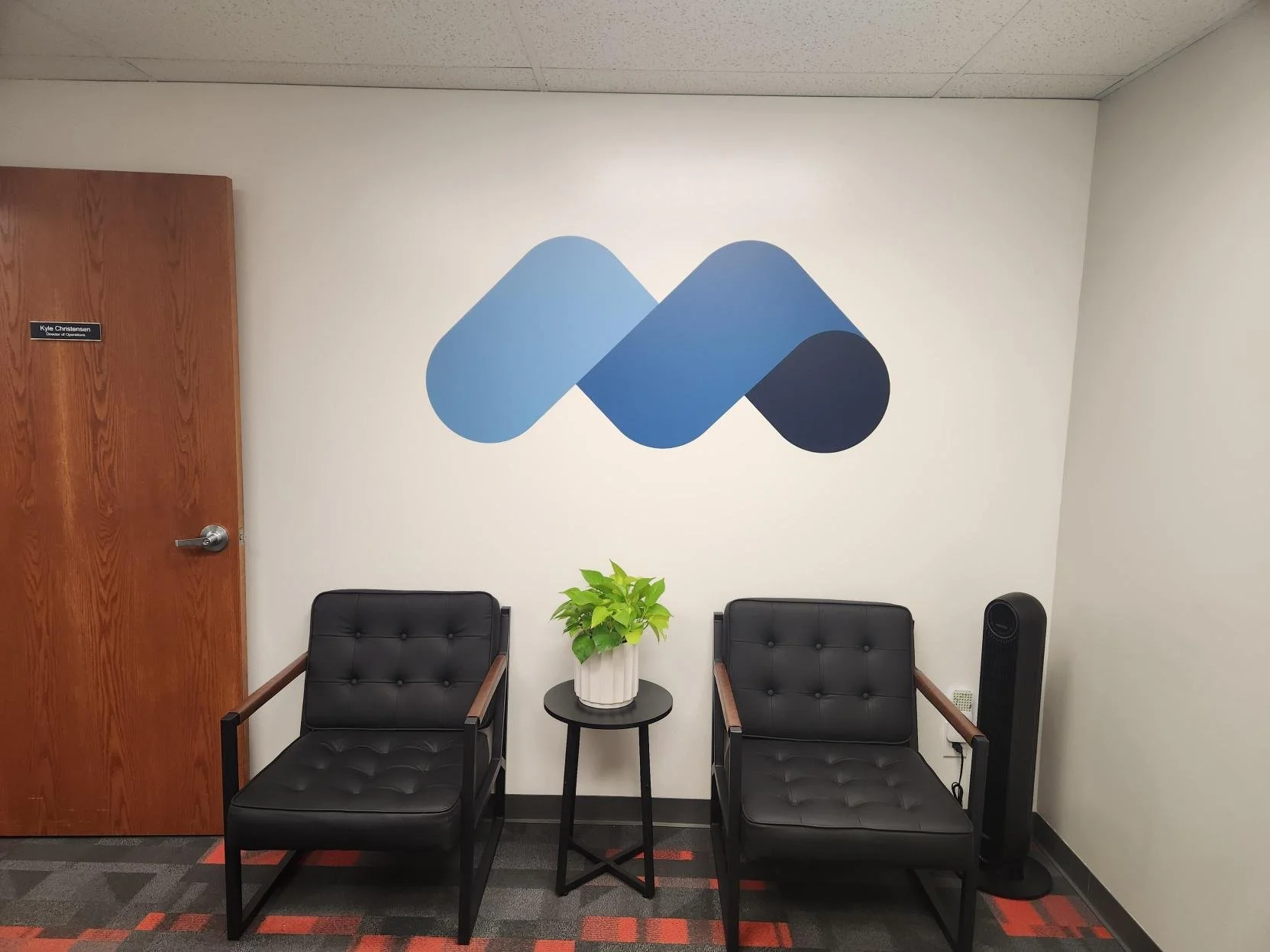 Interior of an office with two black chairs separated by a small black table with a white potted plant, MINDR blue logo on the wall, a wooden door on the left, and black electronic device on the right.