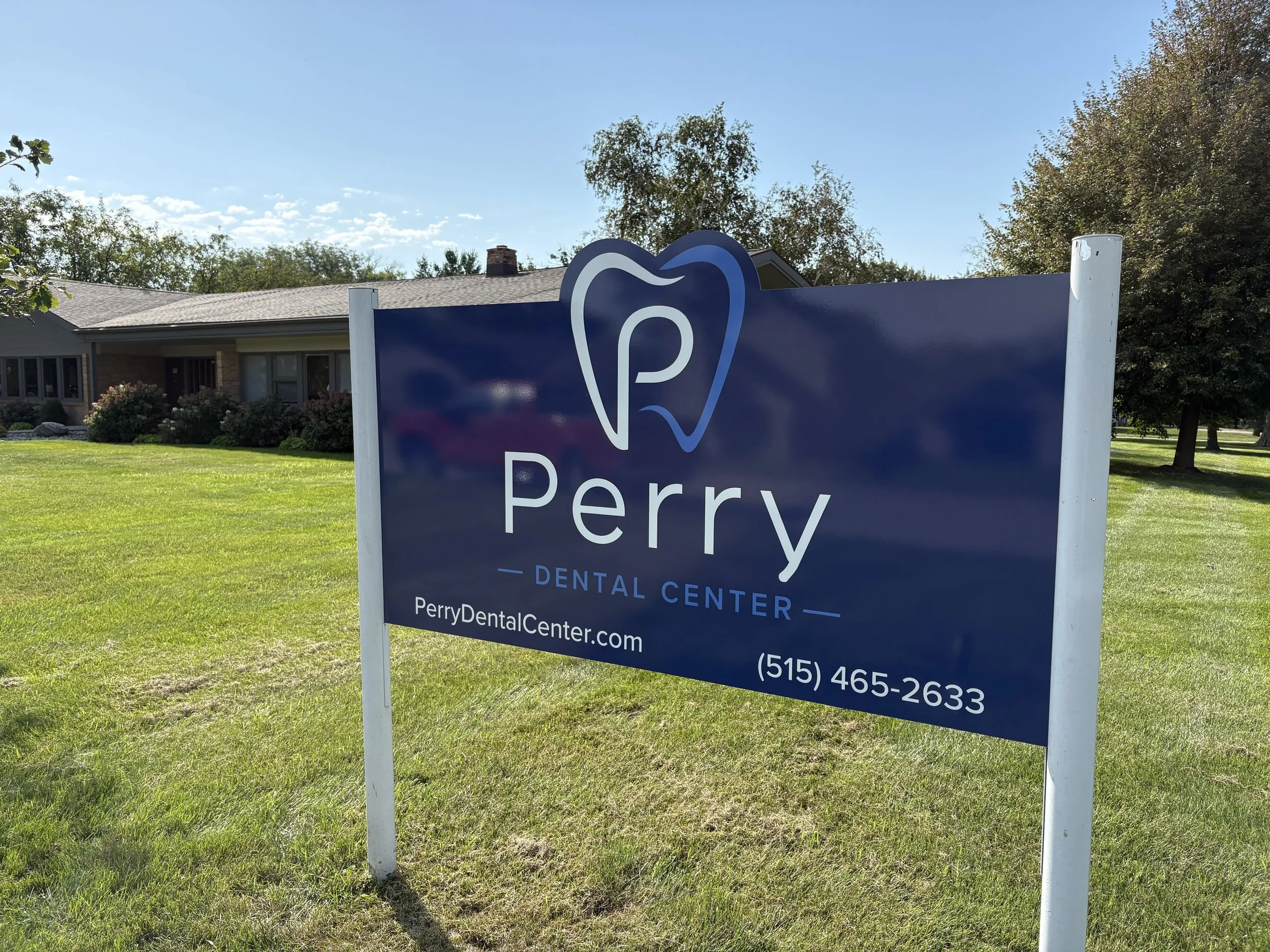Sign for Perry Dental Center with logo of a tooth with a dental tool, located in front of a residential building and trees on a sunny day.