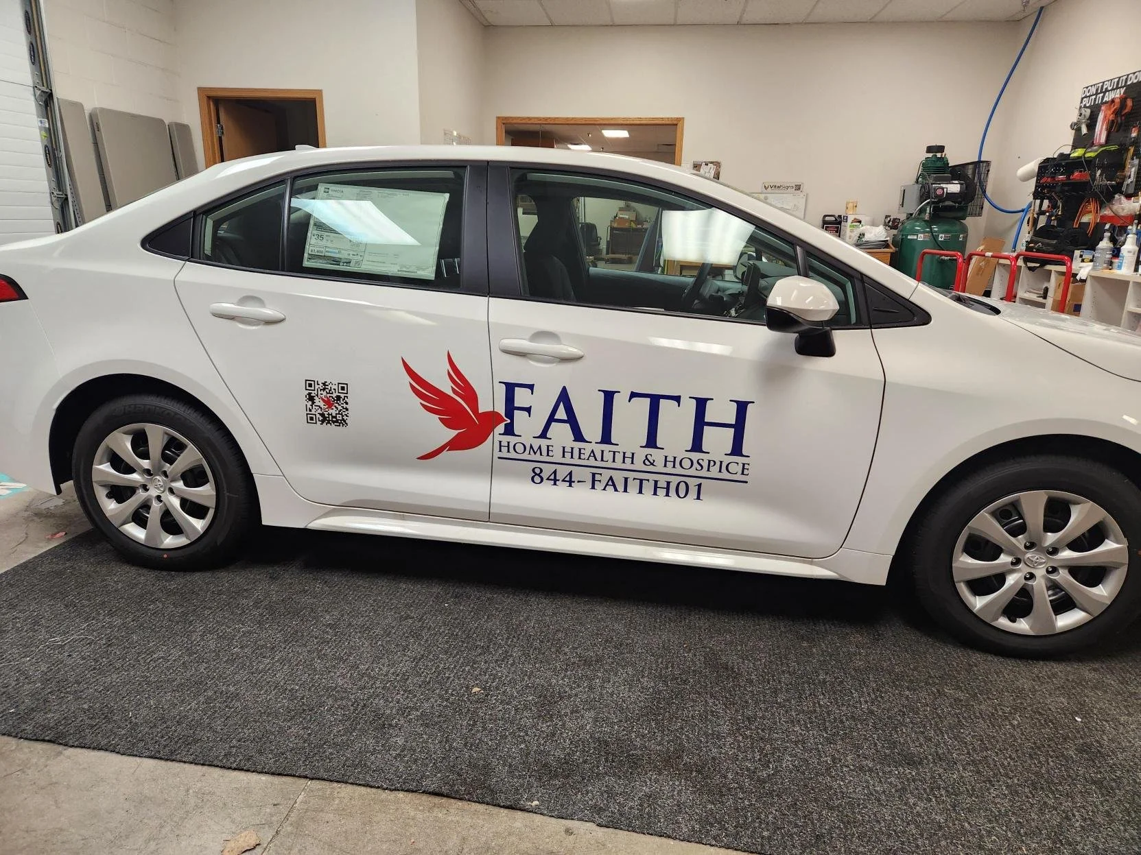 White car with Faith Home Health & Hospice logo, a red dove, and a QR code on the side, parked indoors in a garage or workshop area.