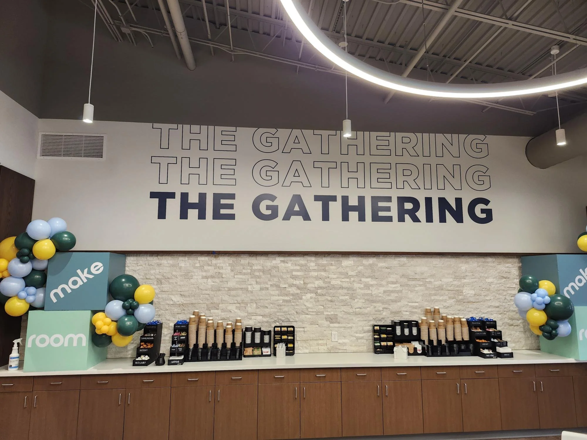 Indoor retail or café space featuring a wall with large text repeating 'THE GATHERING' and 'THE GATHERING' in outlined letters. Decorated with cluster balloons in shades of green, yellow, and light blue on both sides of a green 'make' station. The co