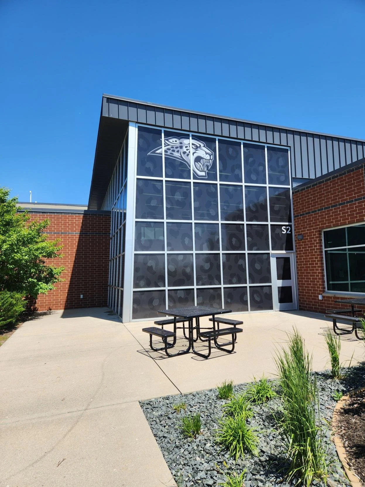 Modern school building with large glass window featuring a jaguar logo and a small outdoor seating area with a picnic table, brick walls, and landscaped greenery under a clear blue sky.