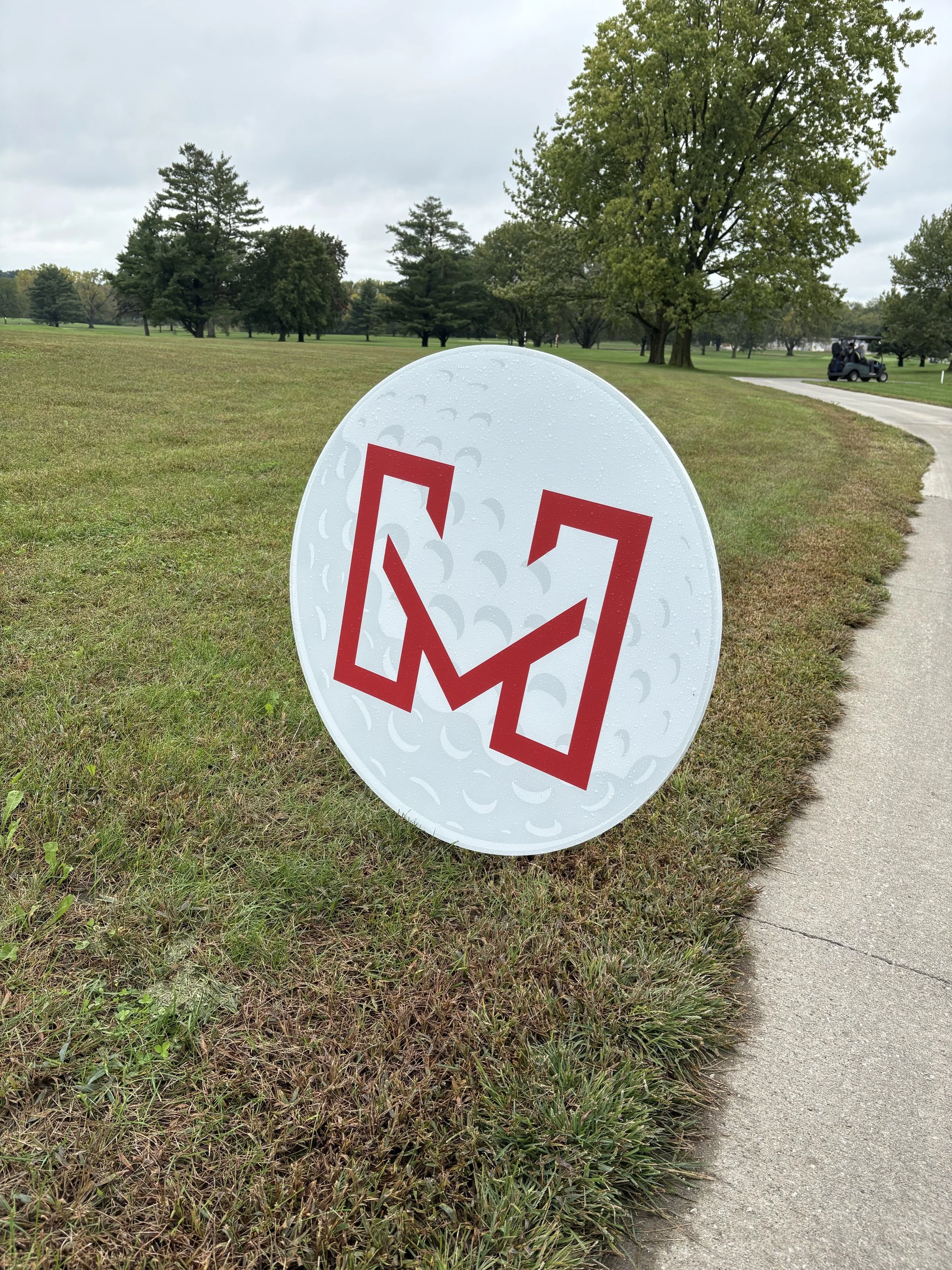 Close-up of a white circular sign with a red slash through a beer mug icon, on a grassy area next to a paved walkway, with trees and a golf cart in the background on a cloudy day.