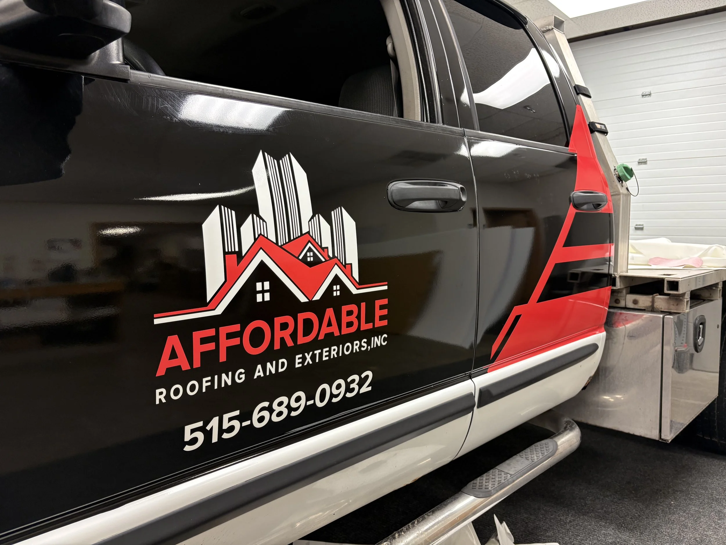 A black service van with a logo featuring a house with a roof, chimney, and multiple windows, and contact info in white and red text on the side, parked indoors next to a garage door.