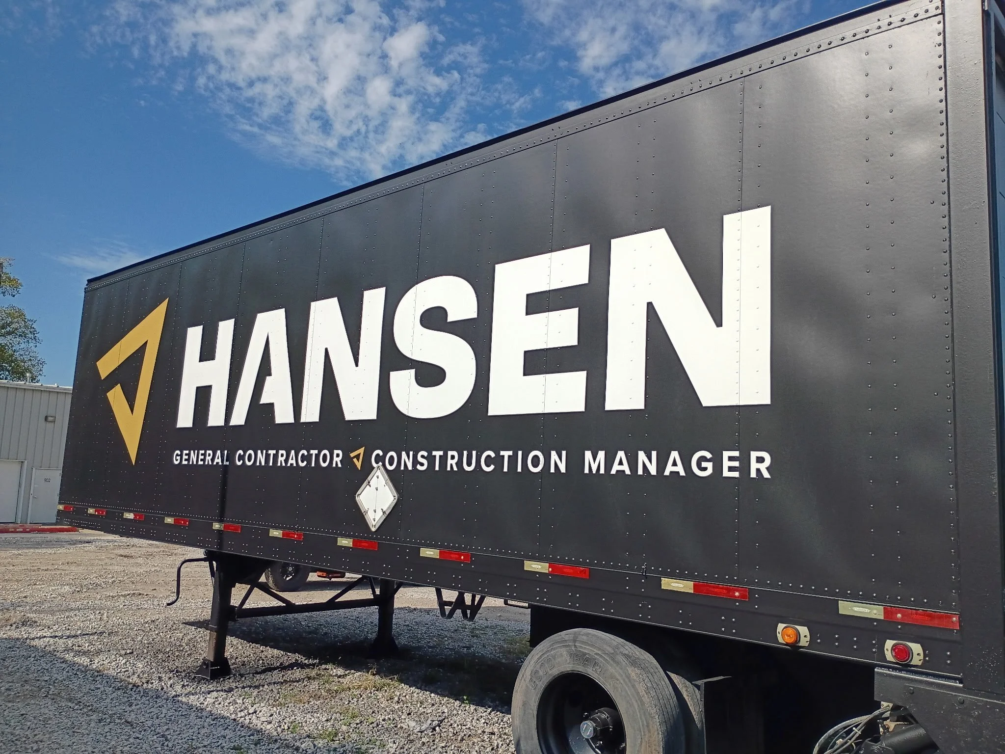 Black trailer with the name 'HANSEN' in large white letters, parked outdoors under a partly cloudy sky.