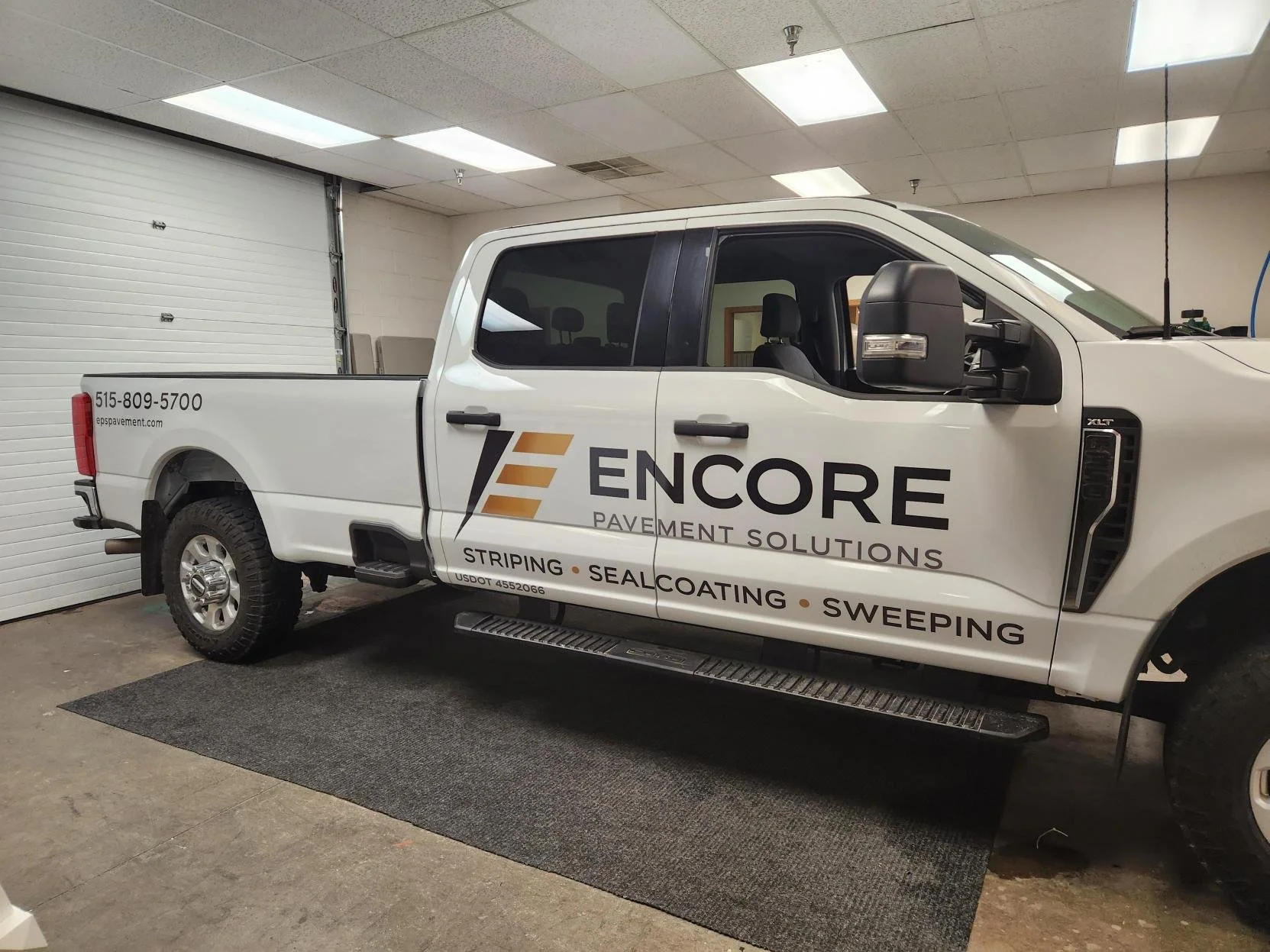 White pickup truck with company logo and contact information parked indoors. The truck advertises Encore Pavement Solutions, which offers striping, sealcoating, and sweeping services.