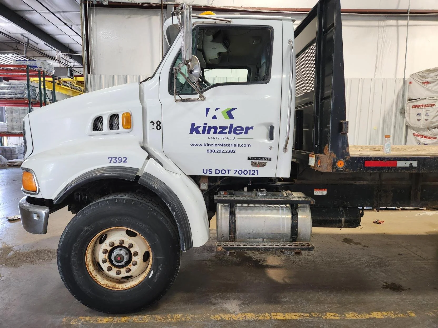 Side view of a white flatbed truck inside a warehouse with company logo and contact information on the door, featuring a large front tire and a rusty rear platform loaded with bags of materials.