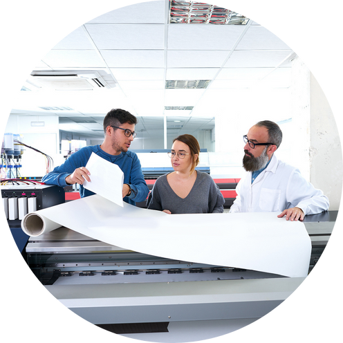 Three people working together in a printing or scanning room, examining large printed material at a machine."}**The image shows three people in a modern, well-lit printing or scanning room. They are gathered around a large format printer or scanner, reviewing a large sheet of printed material.