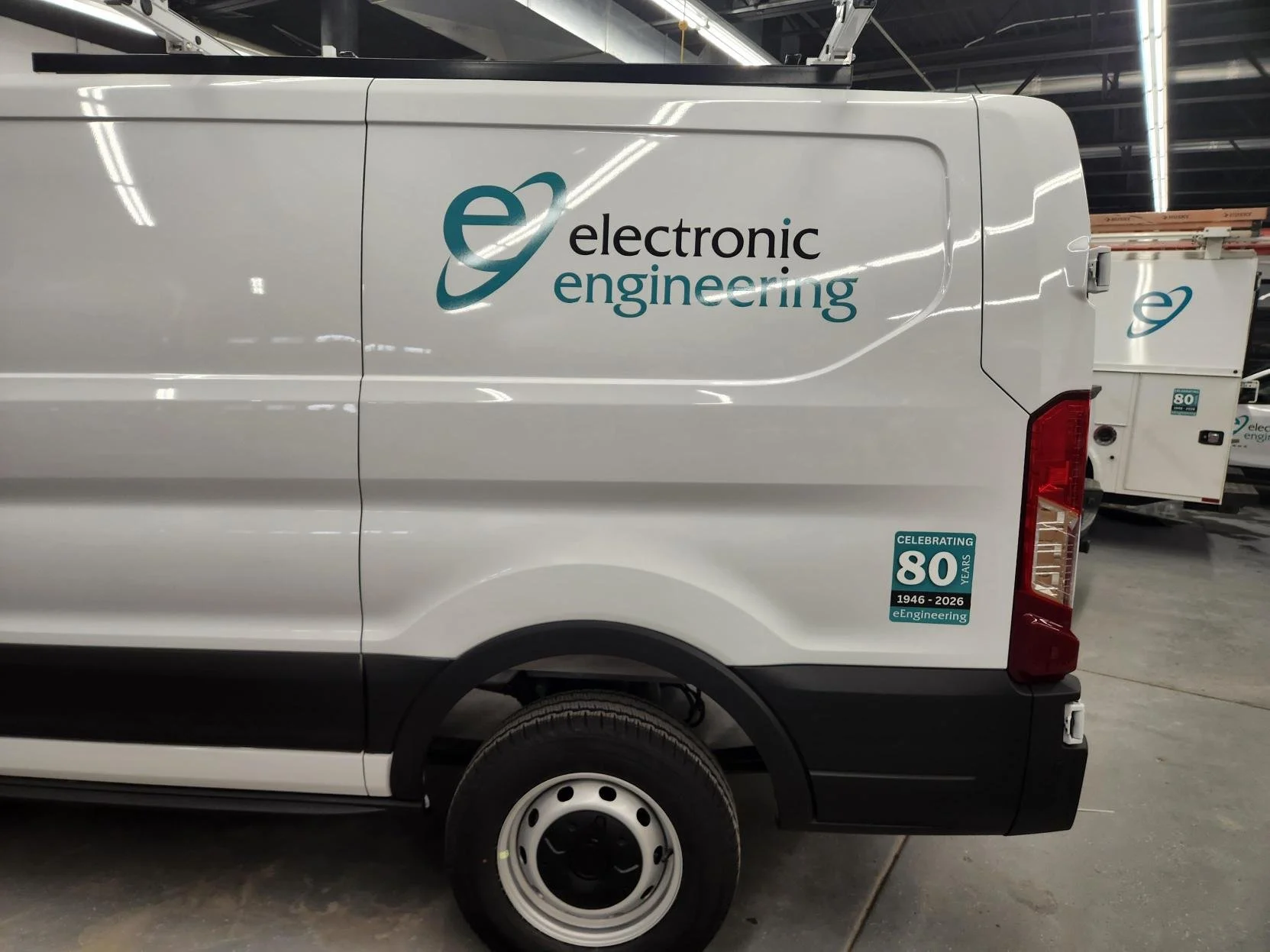 White utility van with electronics engineering logo and a sticker commemorating 80 years of eEngineering on the rear side of the vehicle, inside a warehouse or factory.