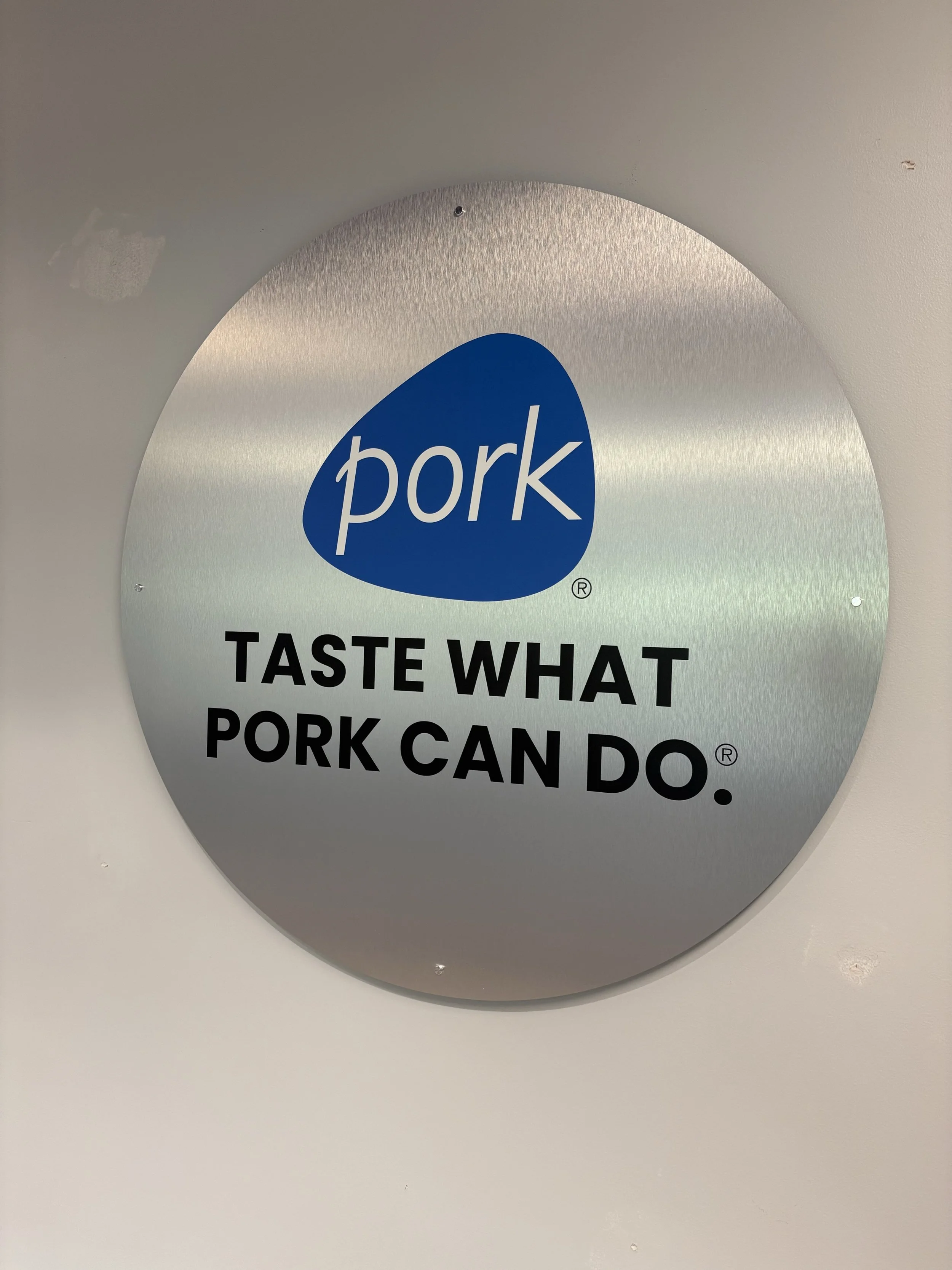 Close-up of a metallic sign with the word 'pork' in white text on a blue background and black text below that reads 'TASTE WHAT PORK CAN DO'.