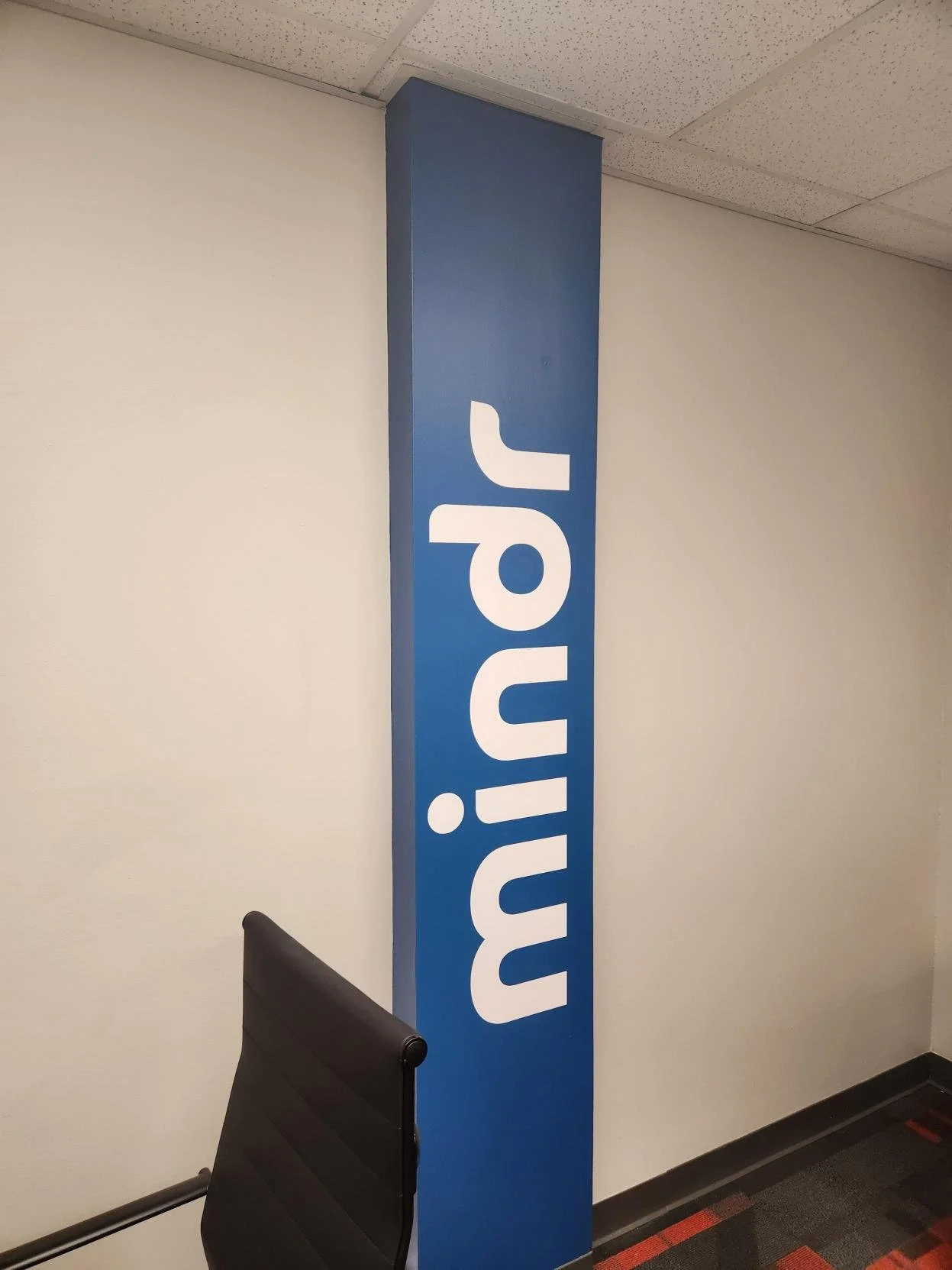 Blue vertical sign with white text spelling 'MINDR' in a corridor with white walls, a black chair, and a carpeted floor.