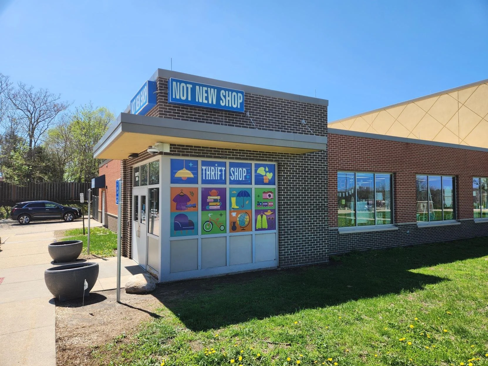 A thrift shop building with a sign that says 'NOT NEW SHOP'. The storefront has colorful icons representing various items like clothes, books, a bicycle, and a globe. The building has large windows and is located in a sunny area with a sidewalk and p
