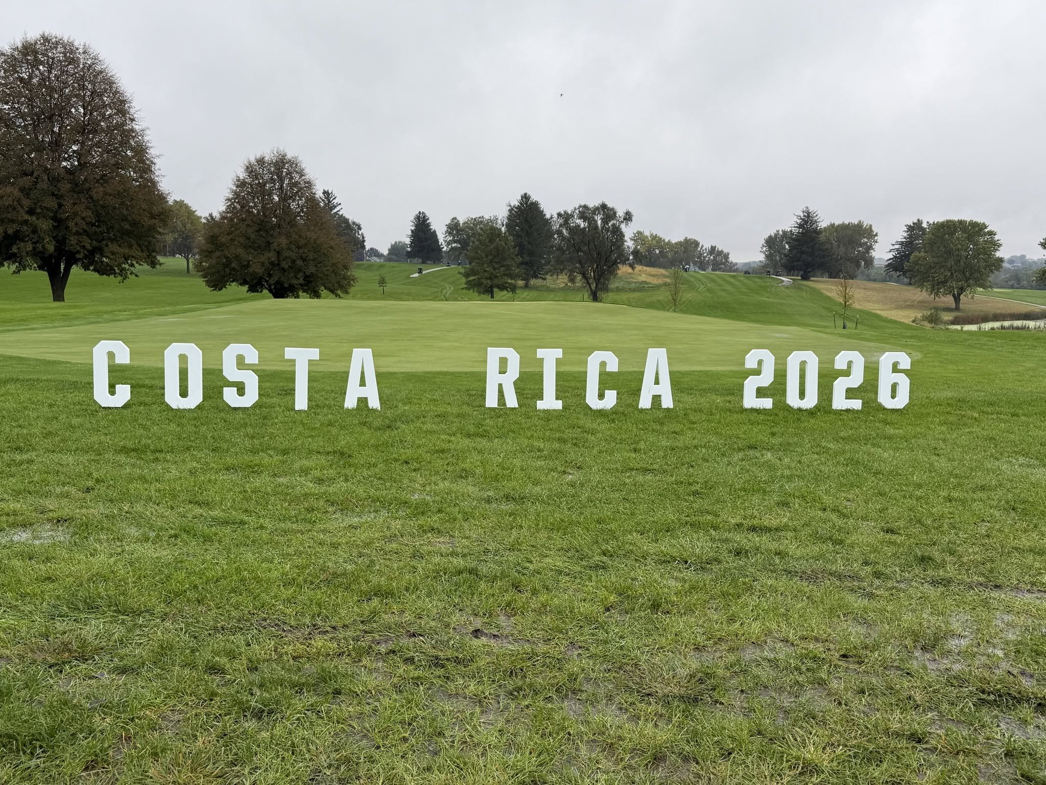 A golf course with a green putting area and trees in the background, under a cloudy sky, with large white letters spelling 'COSTA RICA 2026' on the grass in the foreground.