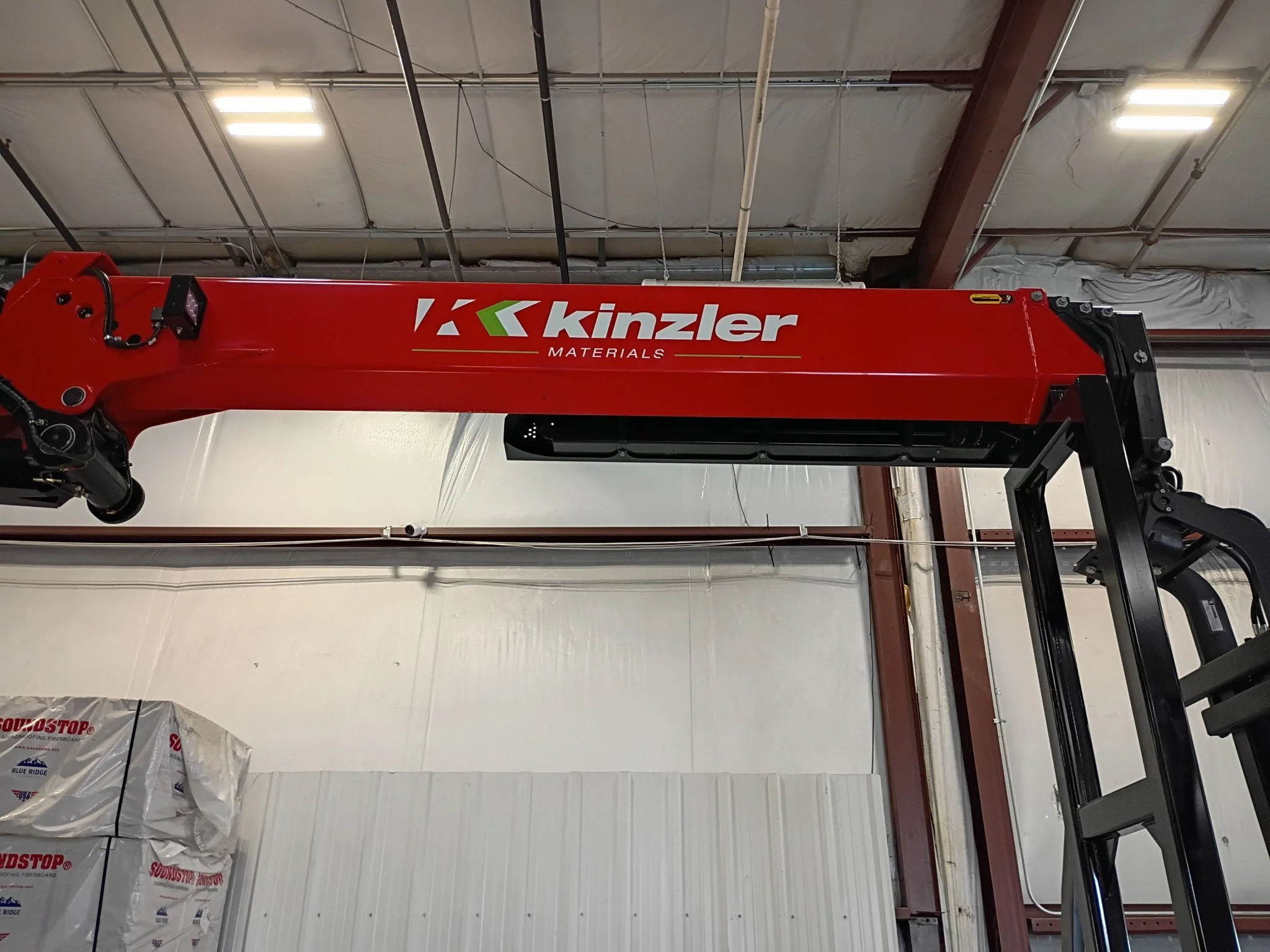 Red Kinzler Materials crane in an industrial warehouse ceiling with pipes and white walls.