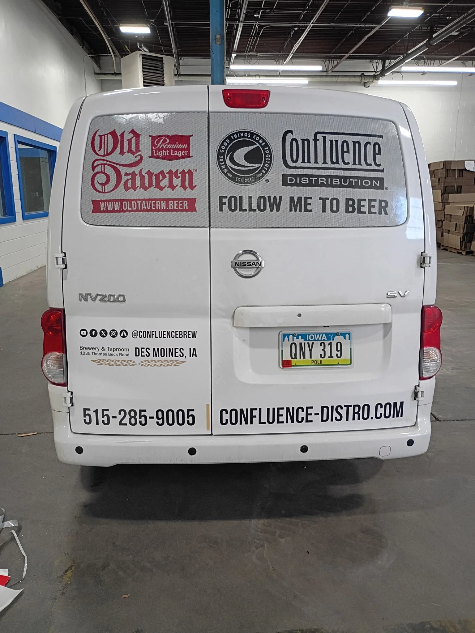 White Nissan NV200 van with logo stickers, text advertising Confluence brew in Des Moines, Iowa, parked in an indoor garage.