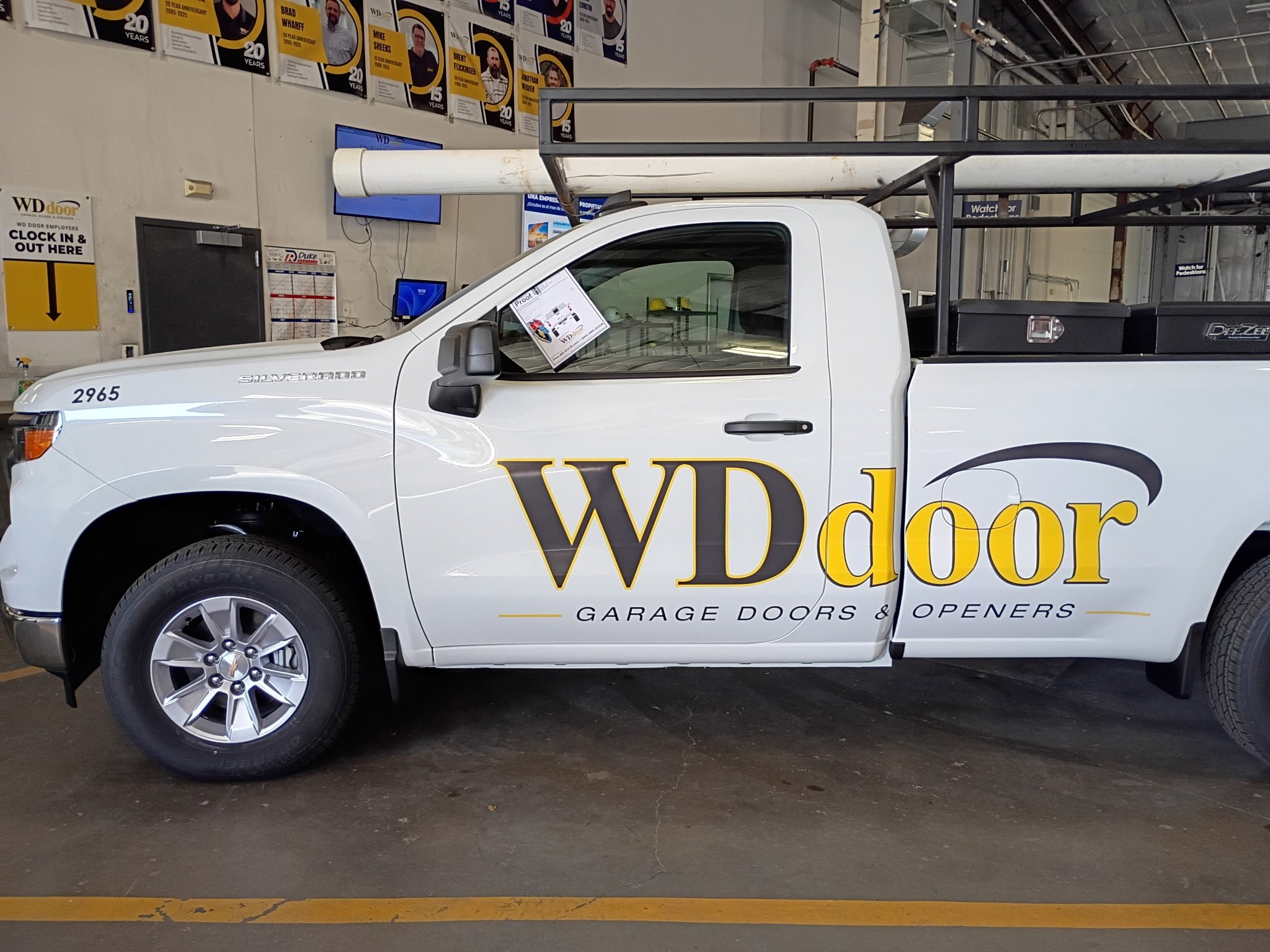 A white pickup truck with black lettering on the side that reads 'WDdoor GARAGE DOORS & OPENERS' parked inside a warehouse or showroom. The truck has a ladder rack on top with a white pipe attached. Behind the truck, there are yellow and purple banne