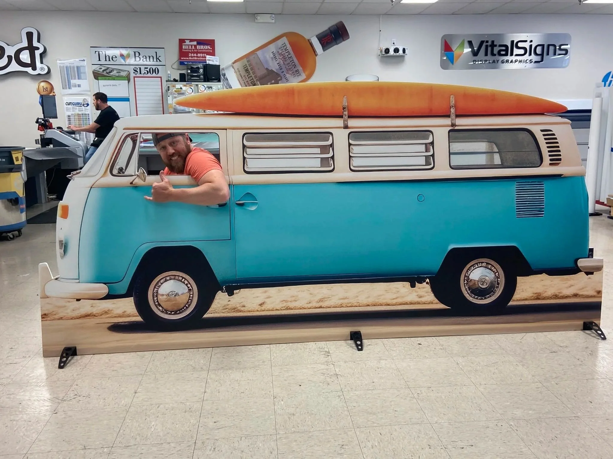 A life-sized display of a vintage Volkswagen van painted in teal and white, with a large orange surfboard on its roof, inside a store with signage and a person working at a checkout counter in the background.