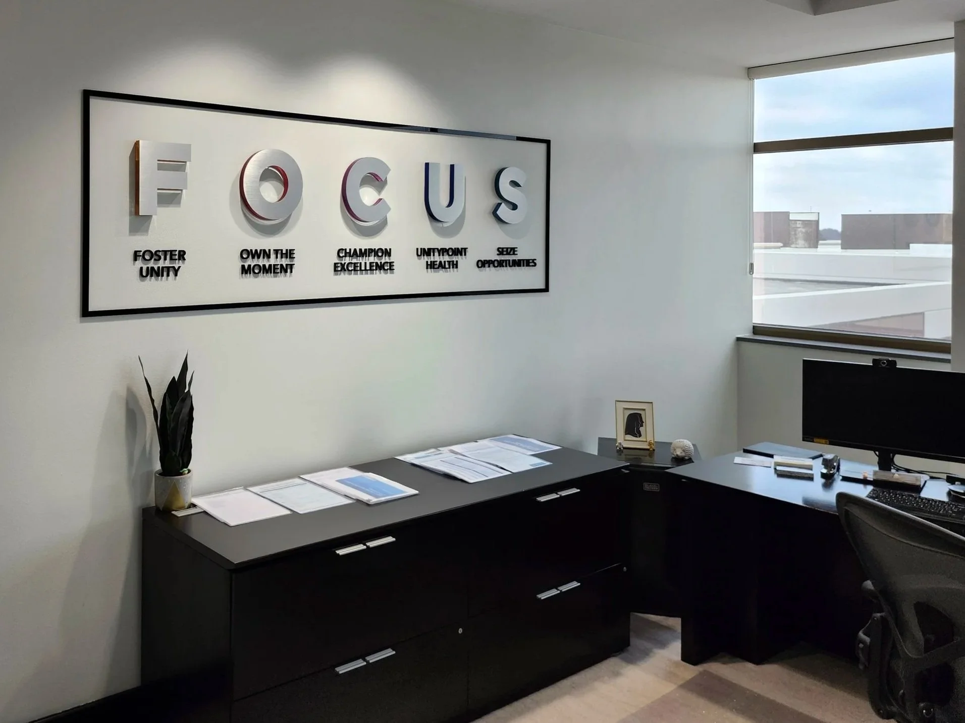 Office with a black desk, computer monitor, and papers; wall art with the word "FOCUS" and motivational phrases underneath; window showing a cityscape.