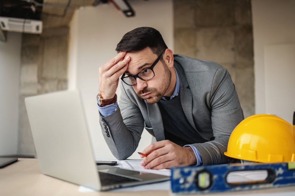 A man in a suit with glasses looks stressed at a laptop, holding his forehead with one hand and a pen in the other. There is a yellow construction helmet on the table.