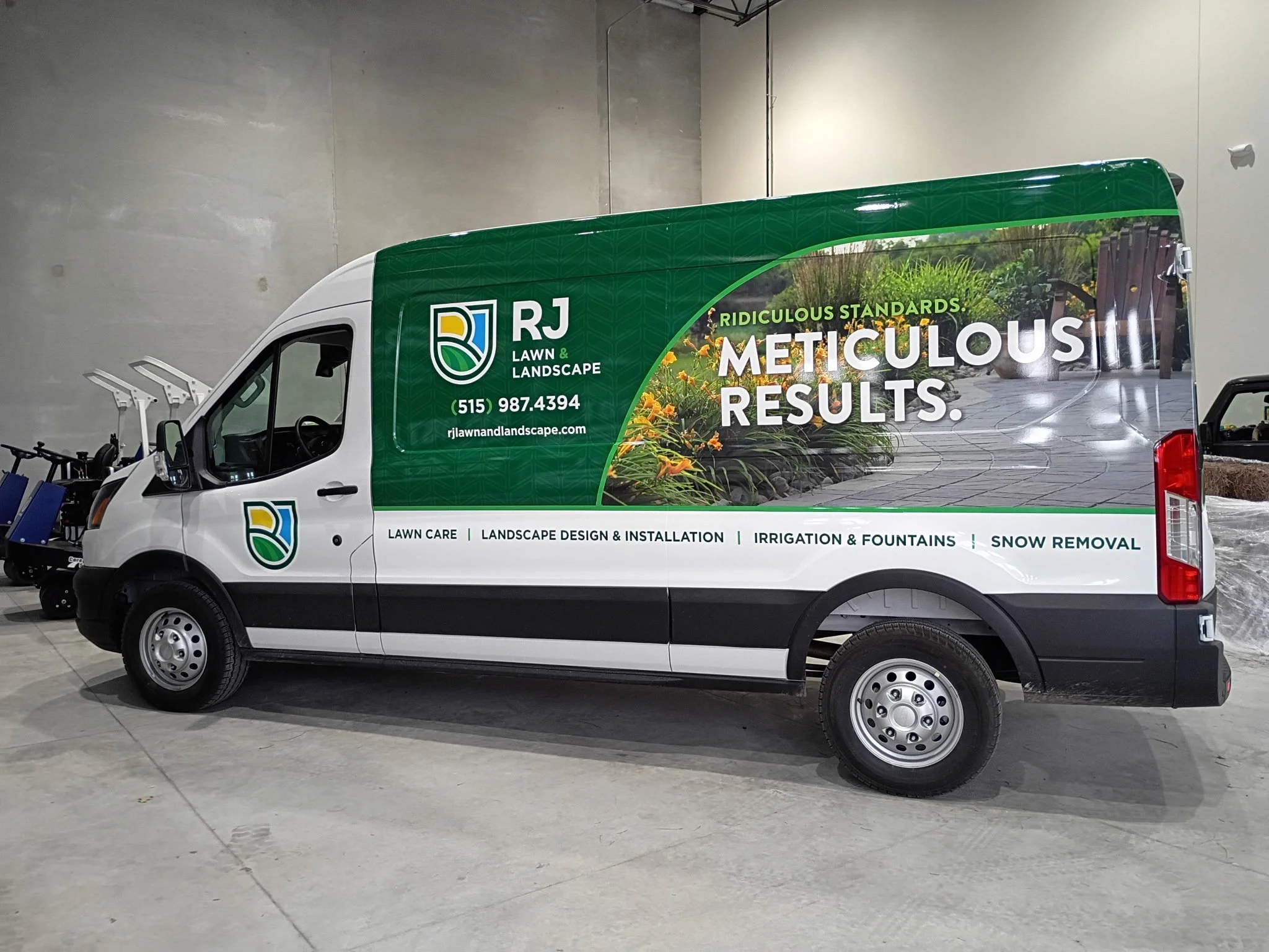 A white delivery van with green marketing graphics parked indoors in a large, empty, concrete-floored space. The van features the logo of RJ Lawn & Landscape, which includes a green and yellow leaf symbol, and has the company's contact number and web