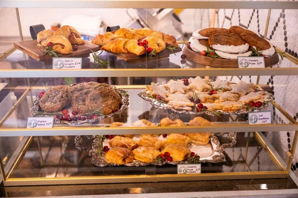 A glass display case filled with baked goods, including cookies, croissants, pies, and cookies decorated for Christmas, with labels and festive decorations.