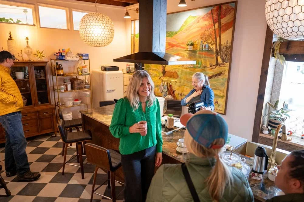 People gathered in a cozy kitchen, smiling and chatting, with a woman in a green shirt in the center holding a drink, a woman in a blue jacket on the counter, and others around, with a large landscape painting on the wall and autumn-themed decorations.