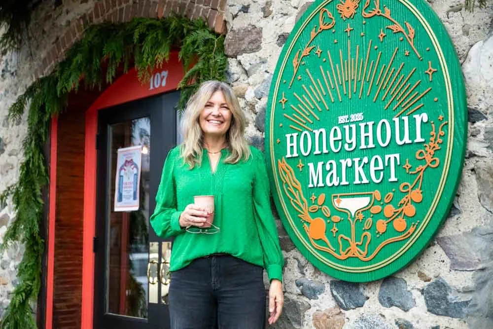 A woman with blonde hair, wearing a bright green blouse and black pants, standing outside the HoneyHour Market, holding a drink in her hand and smiling. The market has a stone wall and a green sign with gold and white lettering.
