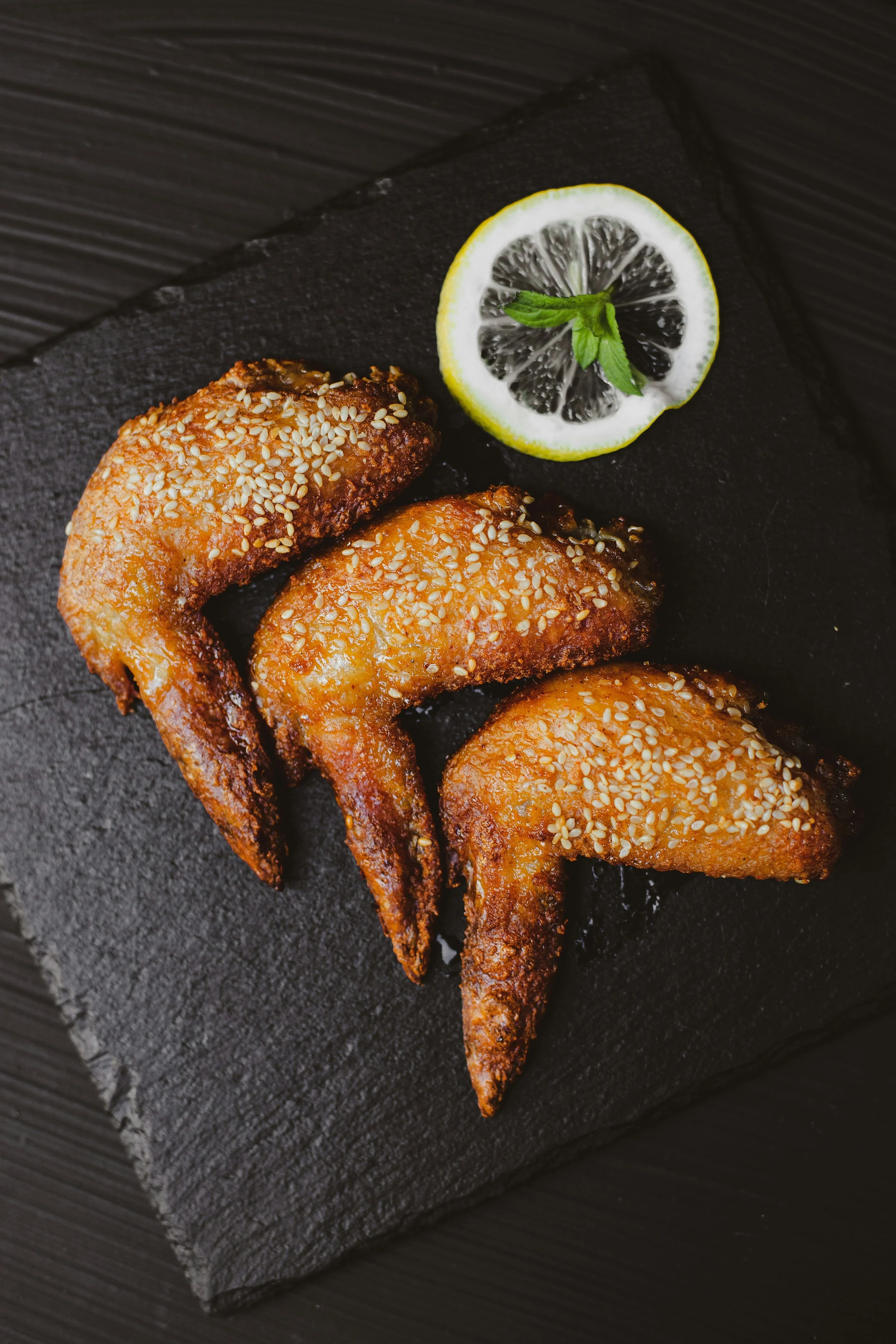 Three sesame seed-topped fried chicken wings on black slate platter, garnished with lemon slice and fresh mint leaf
