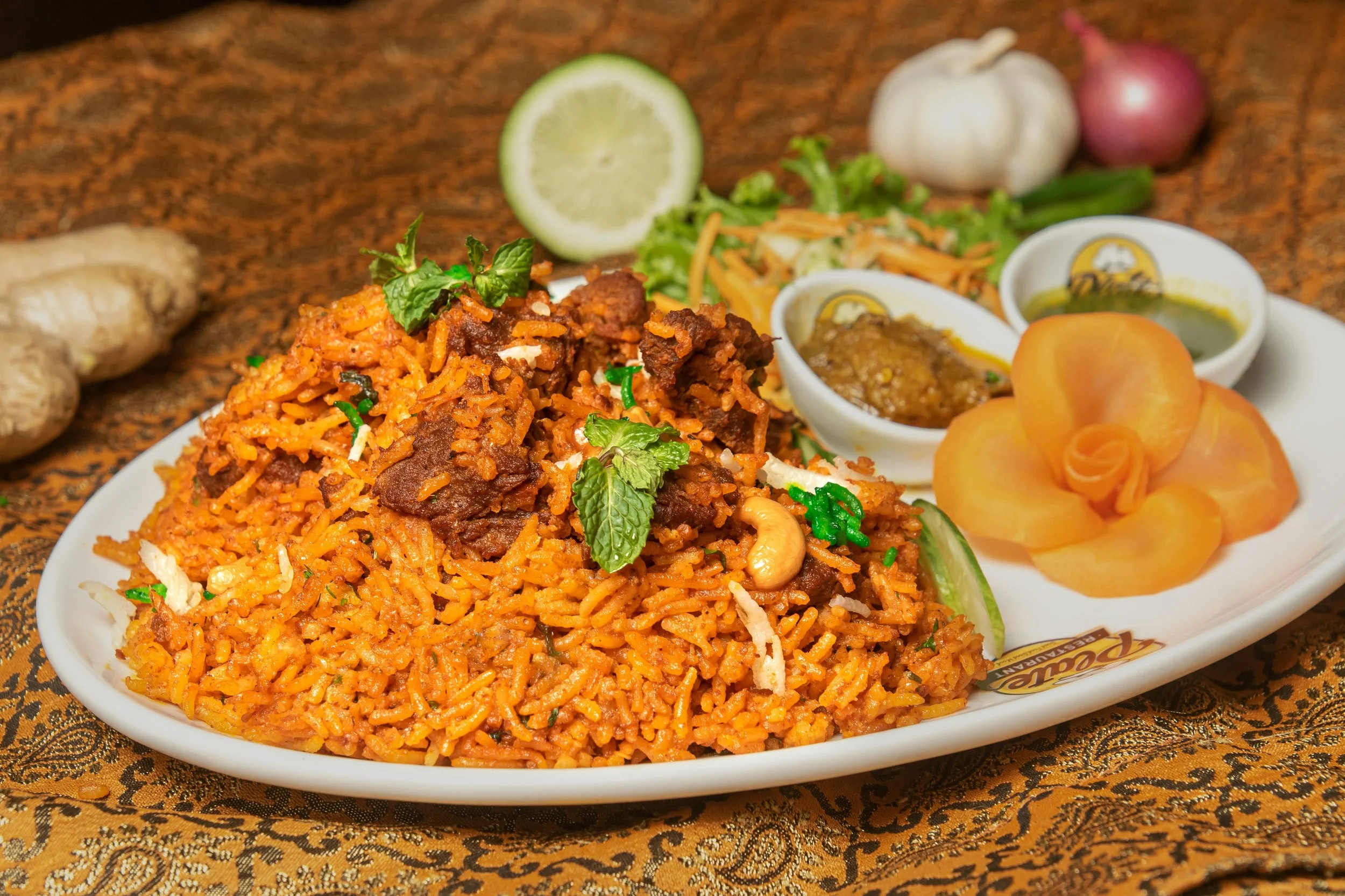 A plate of spicy Indian or Pakistani rice dish with chunks of beef, garnished with fresh cilantro, served alongside small bowls of curry and pickles, with lime, garlic, and onions in the background.
