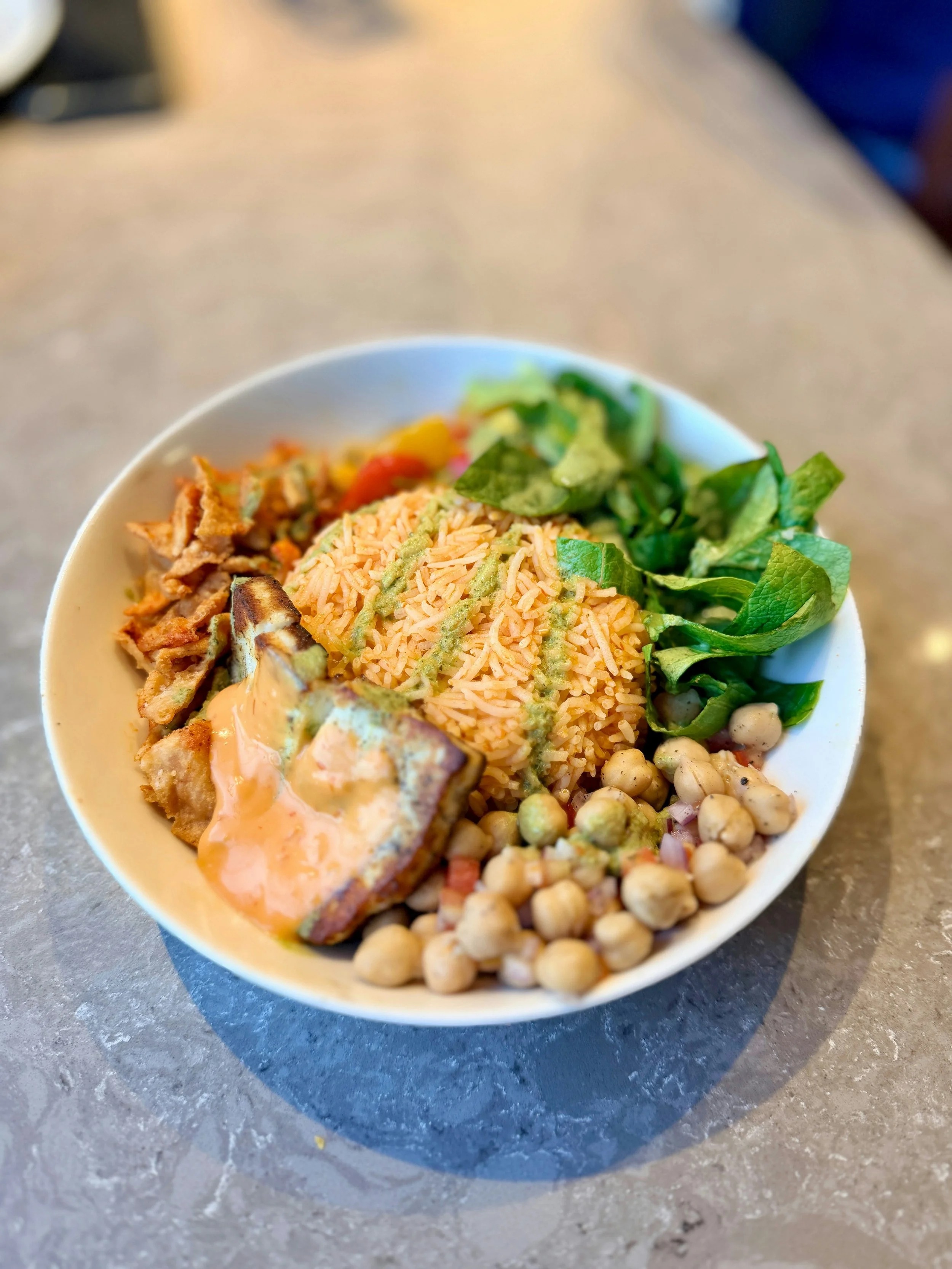 A white bowl filled with rice, chickpeas, chopped vegetables, lettuce, and some cooked meat, placed on a gray marble surface.