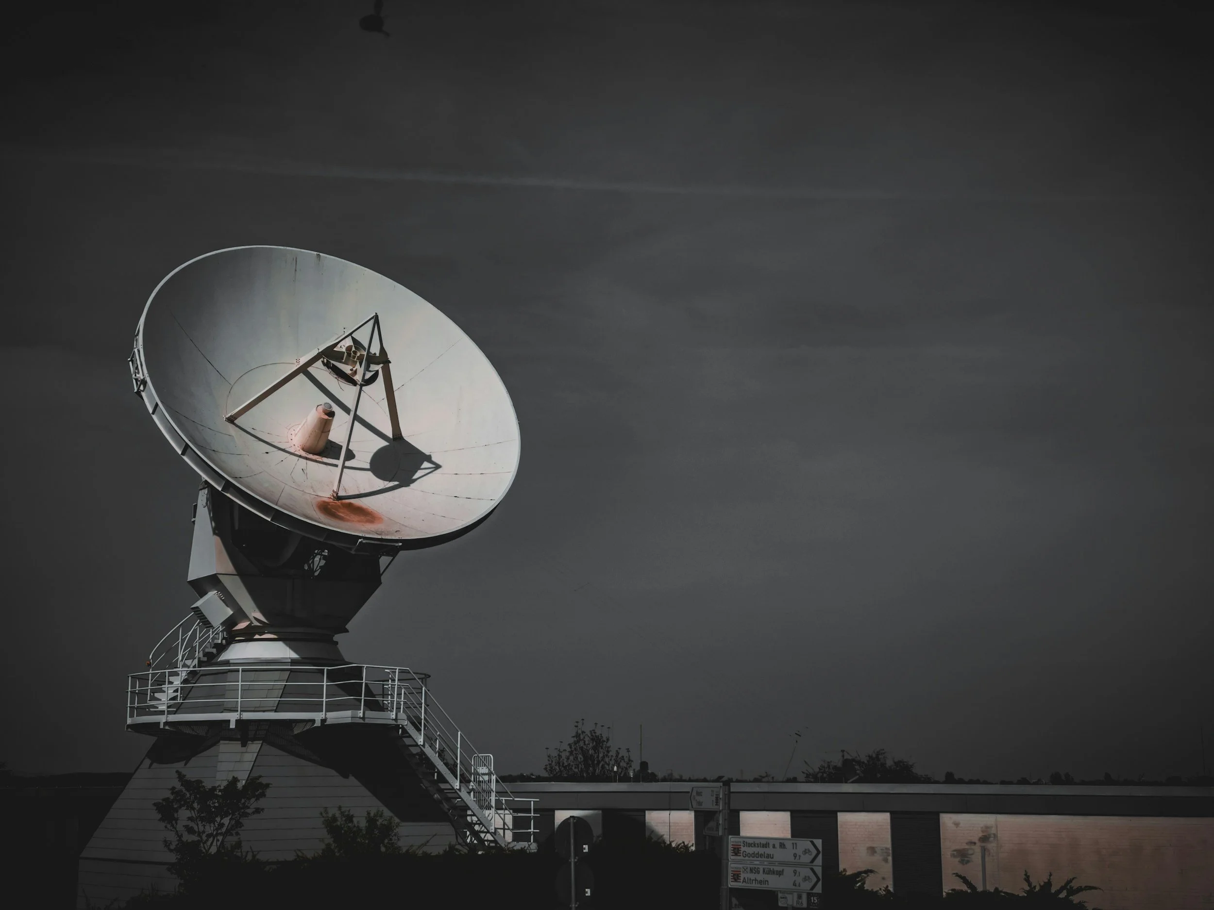 A large satellite dish antenna on a rooftop against a dark, cloudy sky, with a section of outdoor stairs visible below.