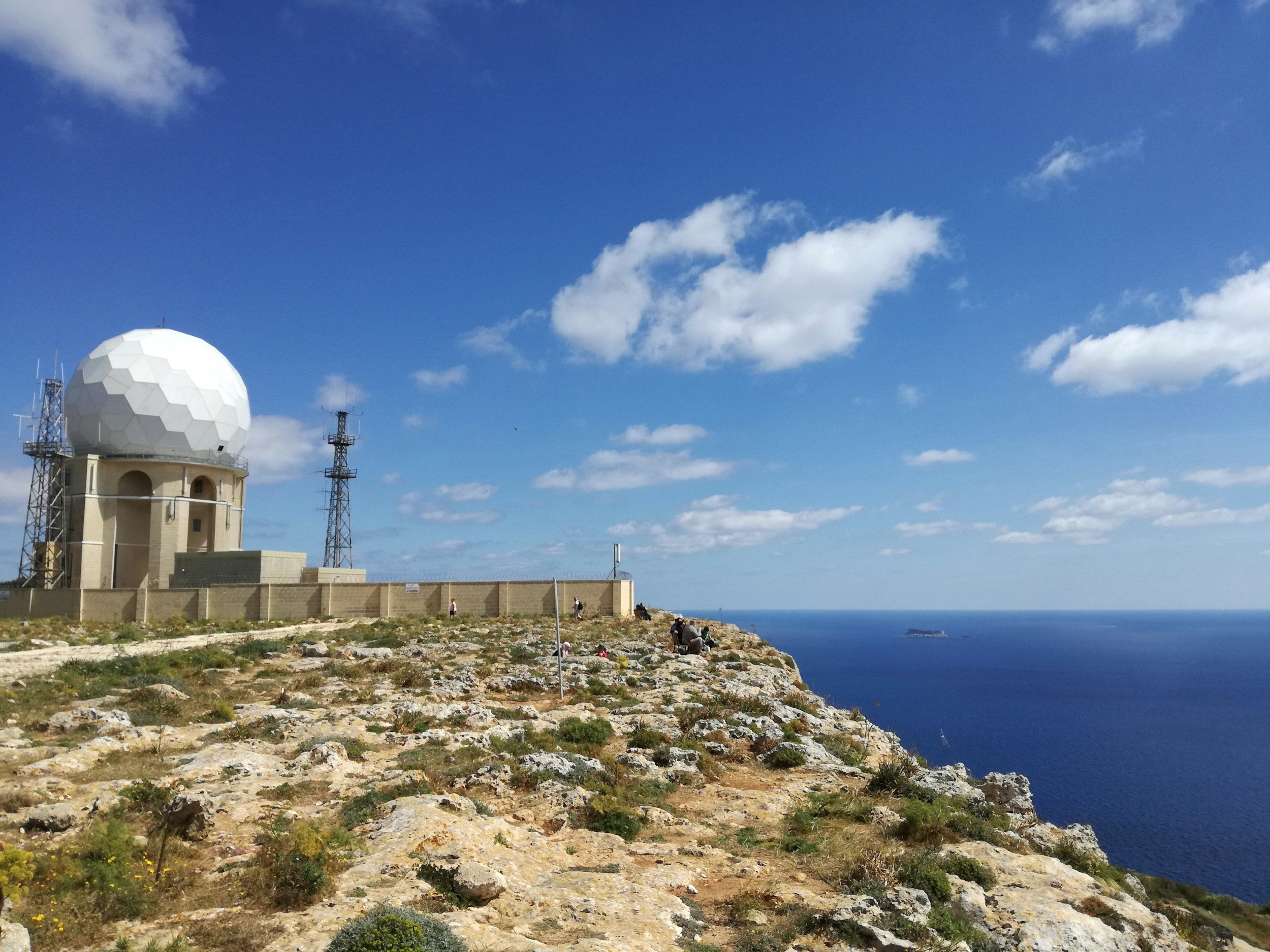 A blue sky with scattered white clouds over a rocky landscape with sparse vegetation, a white dome-shaped building with antennas, and several people sitting or walking along the edge of the rocky area by the sea.
