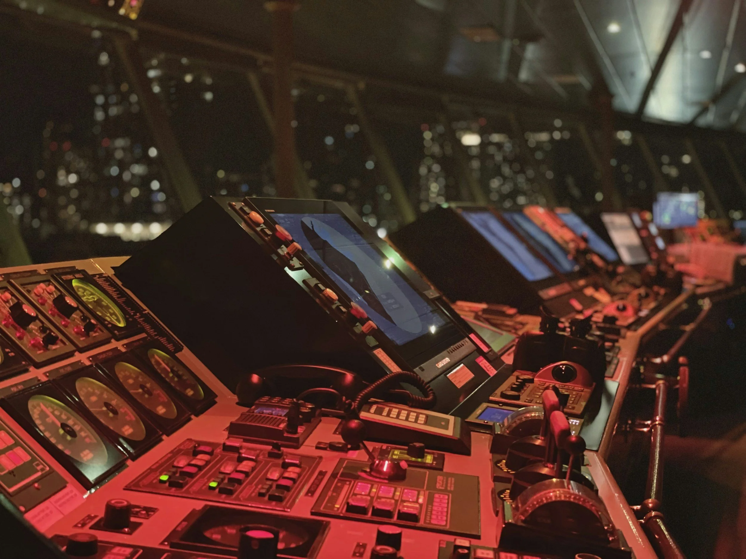 A DJ setup with multiple electronic music equipment and turntables, illuminated with red and orange light, and a dark, blurry background showing an illuminated cityscape at night.