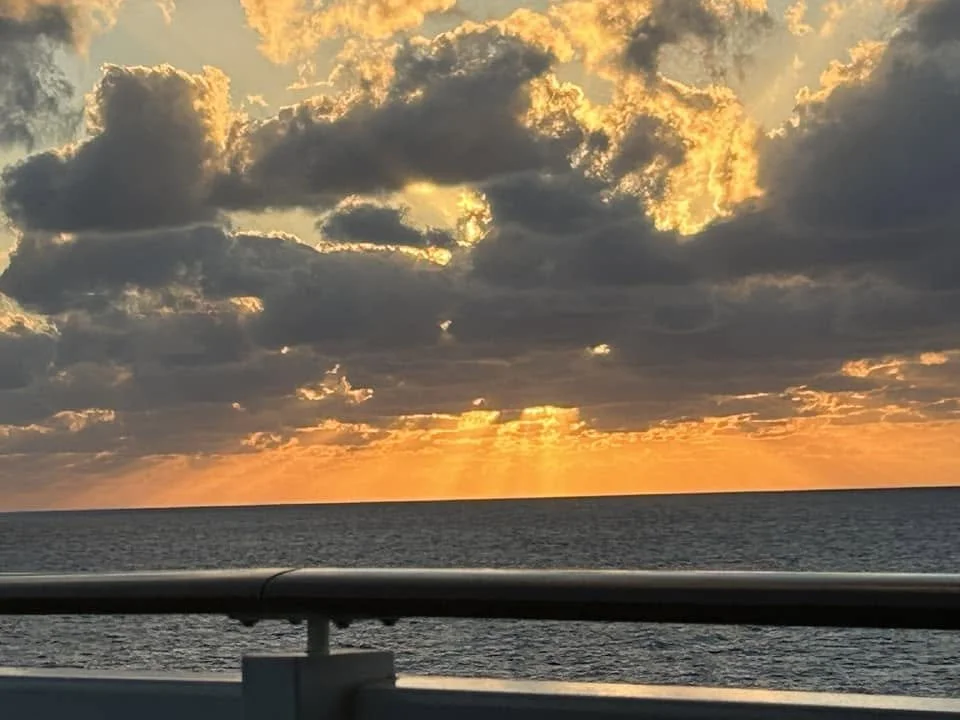 Sunset over the ocean with a cloudy sky, viewed from a boat or bridge railing.