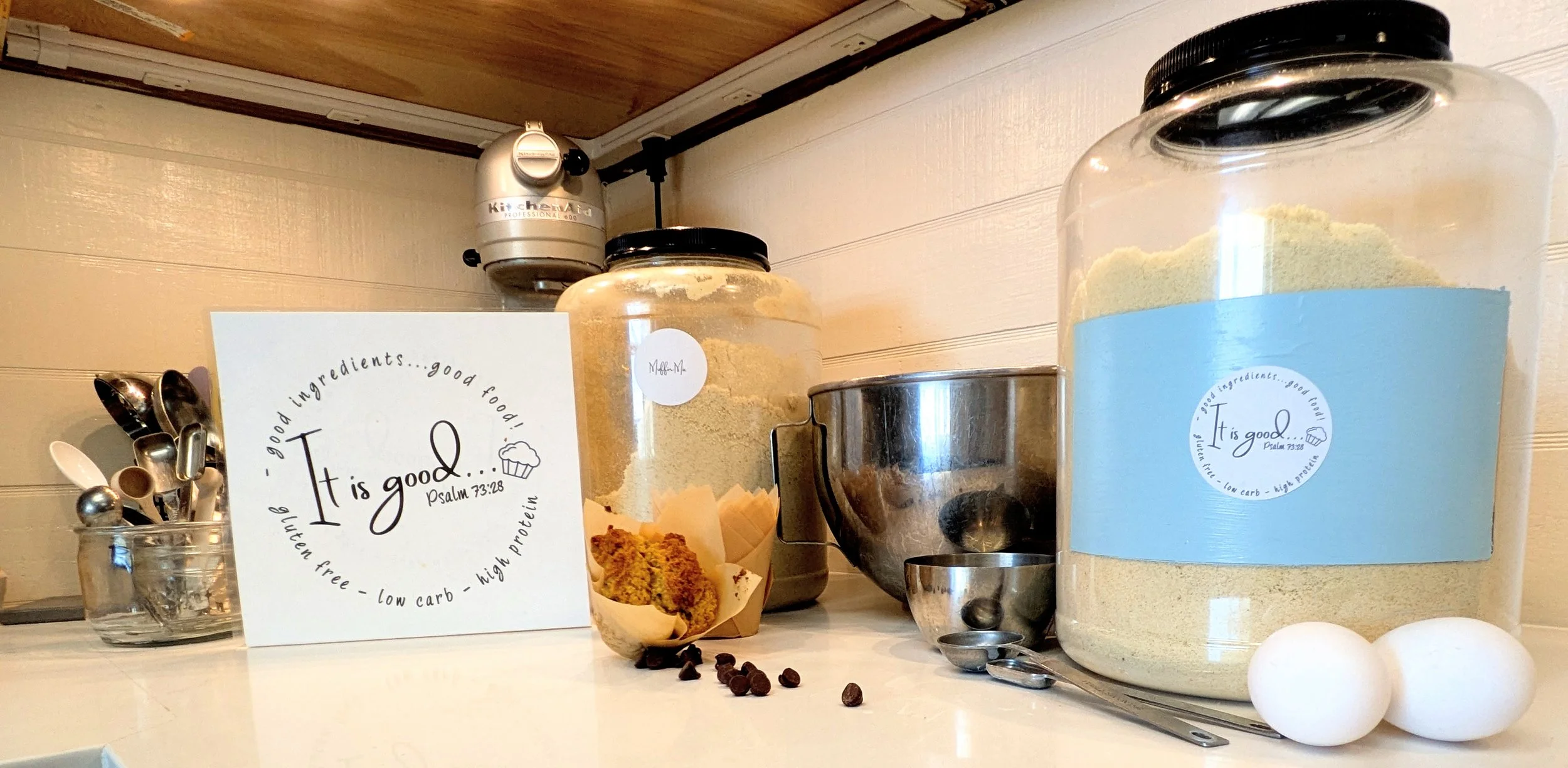 A kitchen counter with various baking ingredients and utensils. There are large containers labeled with a quote from Psalm 73:28, a muffin in paper liner, a small jar of baking powder, metal measuring cups, a metal mixing bowl, and a few eggs. A sign reads, 'It is good,' referencing healthy eating and high protein, low carb baking.